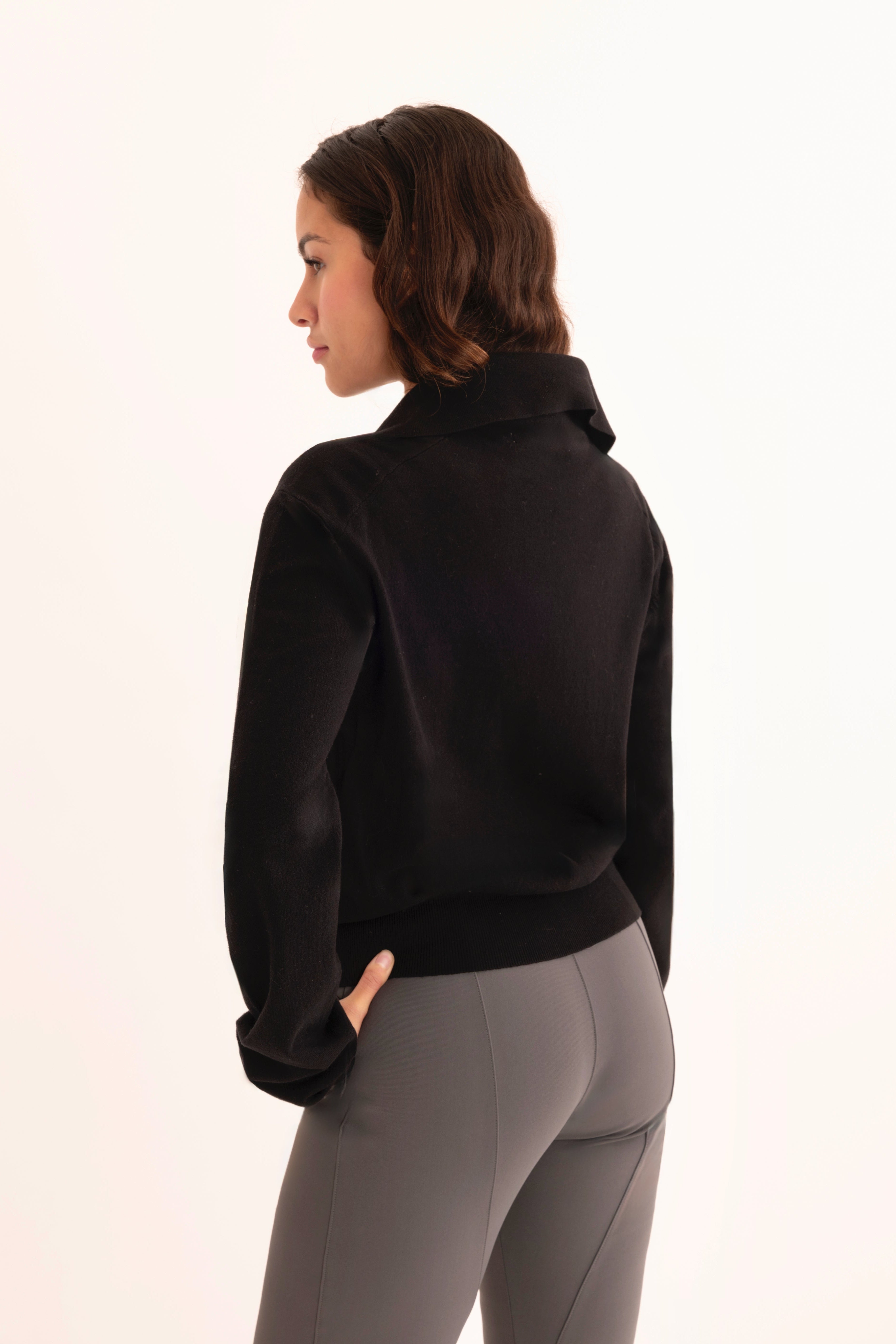 Back view of woman wearing a black zip-up jacket and gray pants on a white background