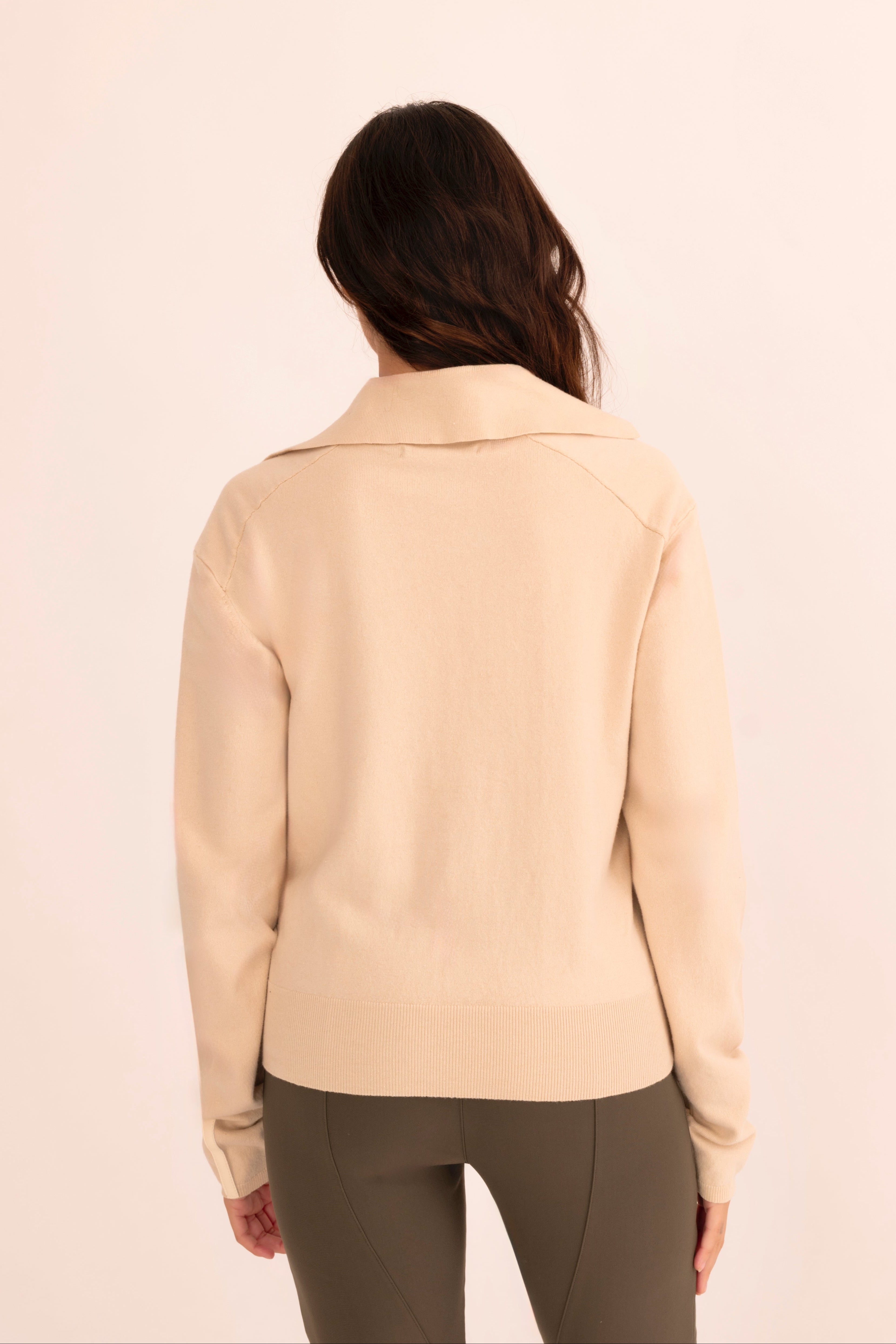 Back view of woman wearing a beige zip-up jacket and green pants on a white background