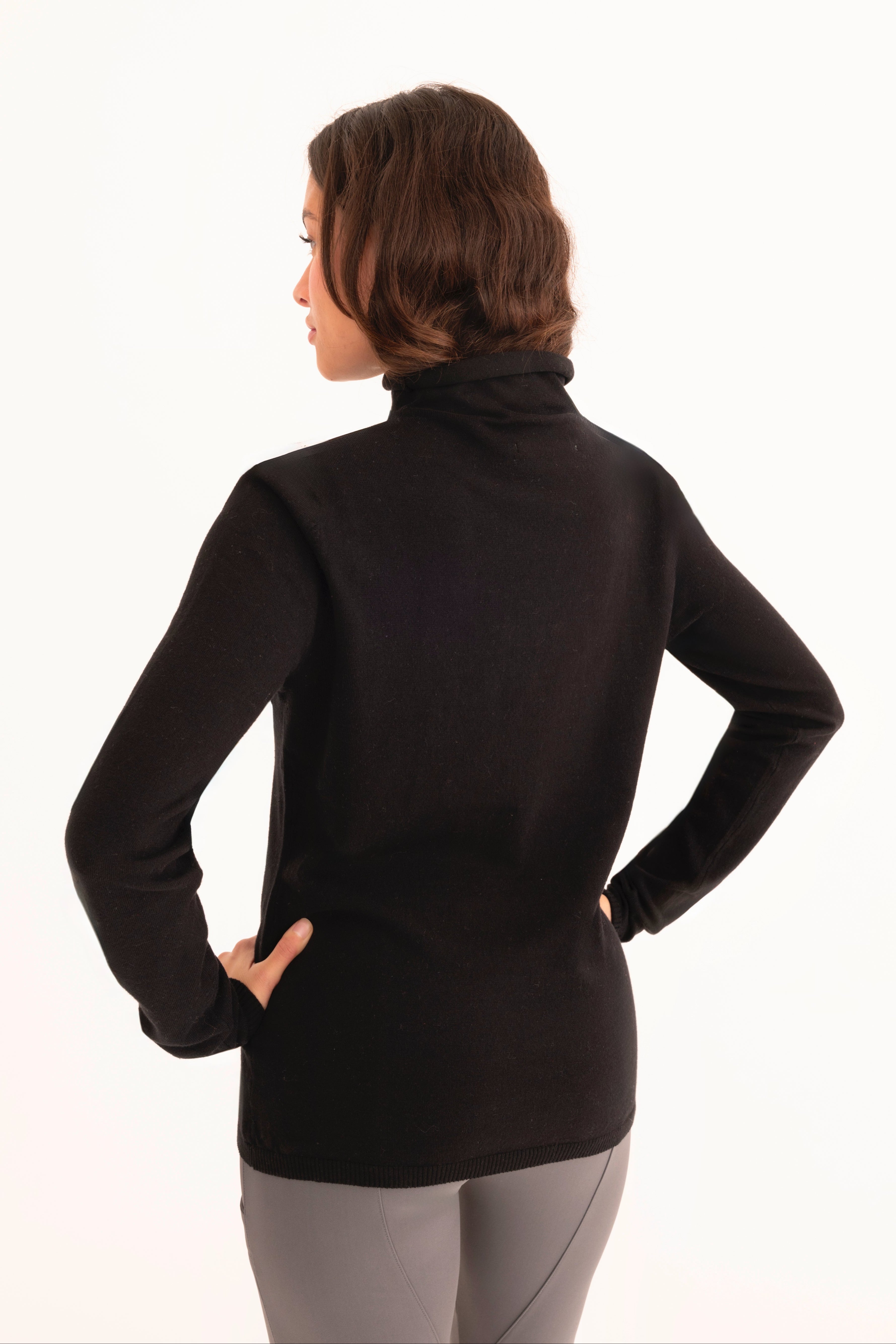 Back view of a woman wearing a black funnel neck top and gray pants on a white background