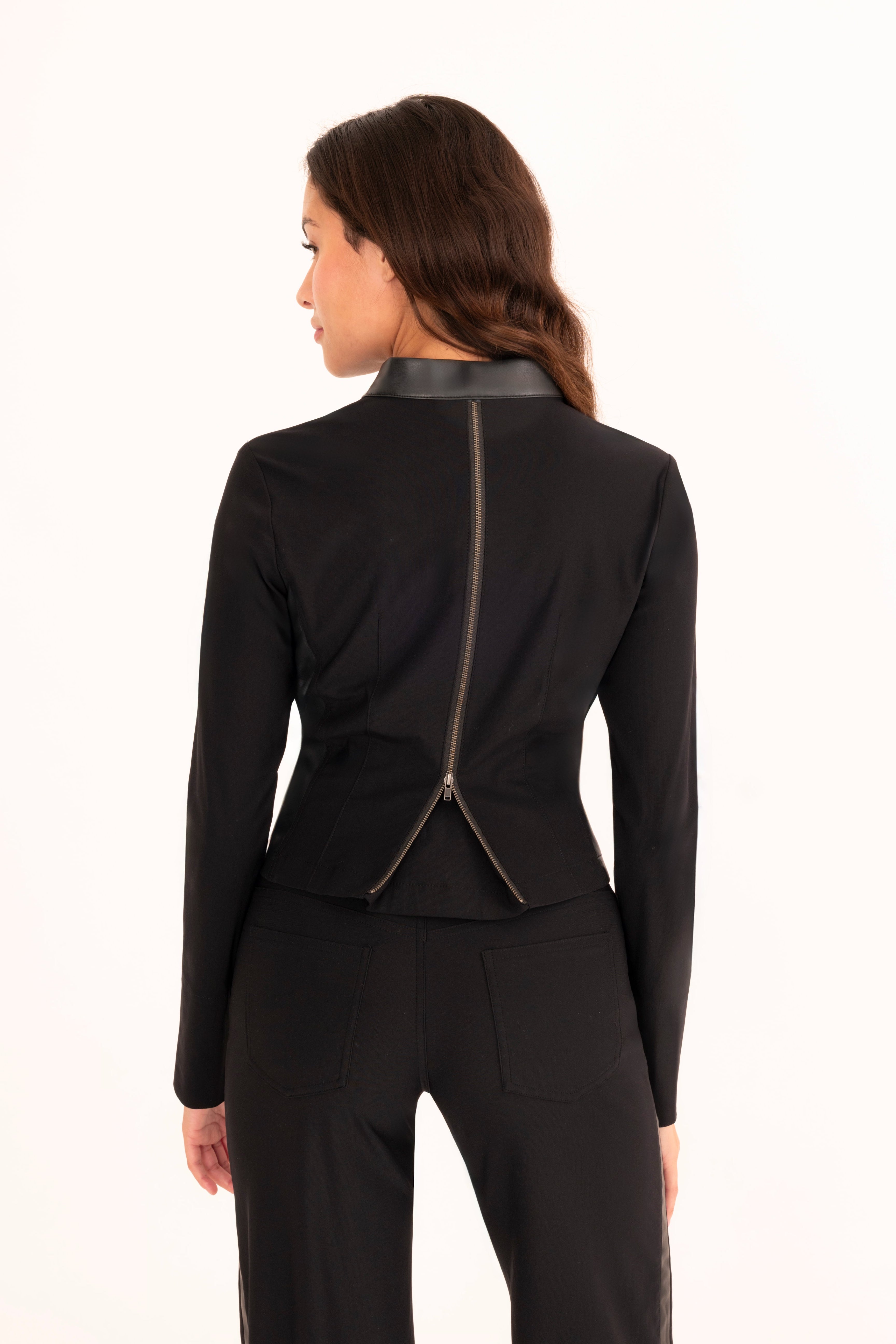 A woman shown from the back wearing a black long-sleeve fitted top with a prominent metallic zipper running down the center back and collar, paired with black pants, against a plain white studio background.