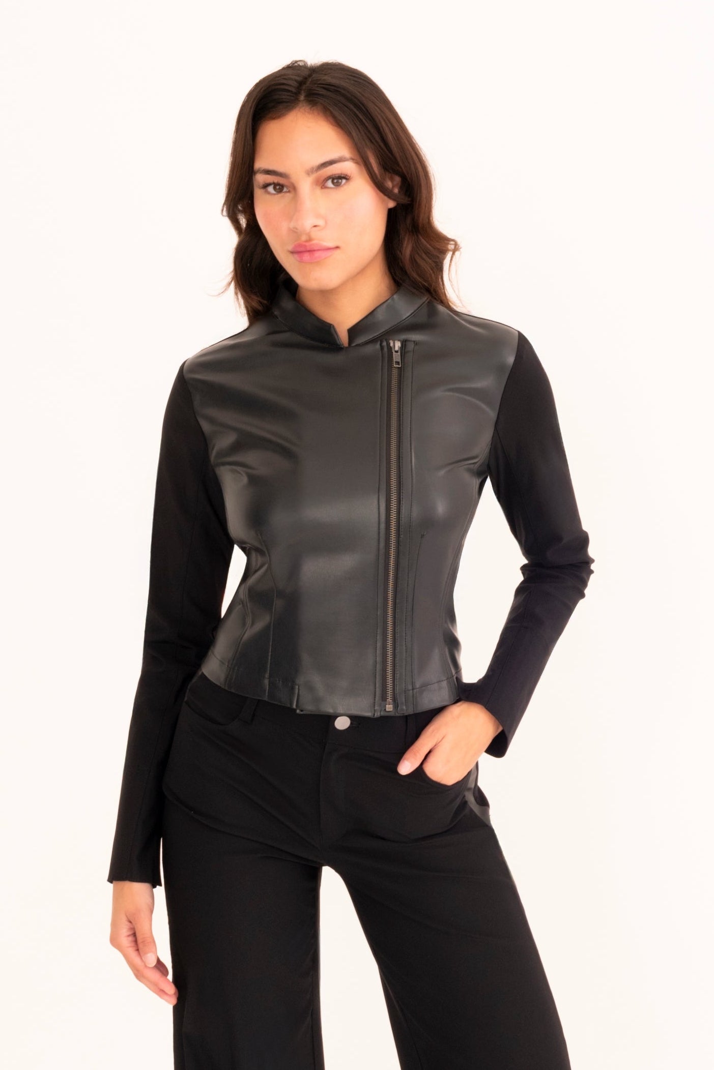 A woman modeling a black faux-leather motorcycle jacket with a stand collar and front zipper, paired with matching black pants, against a plain off-white studio background.