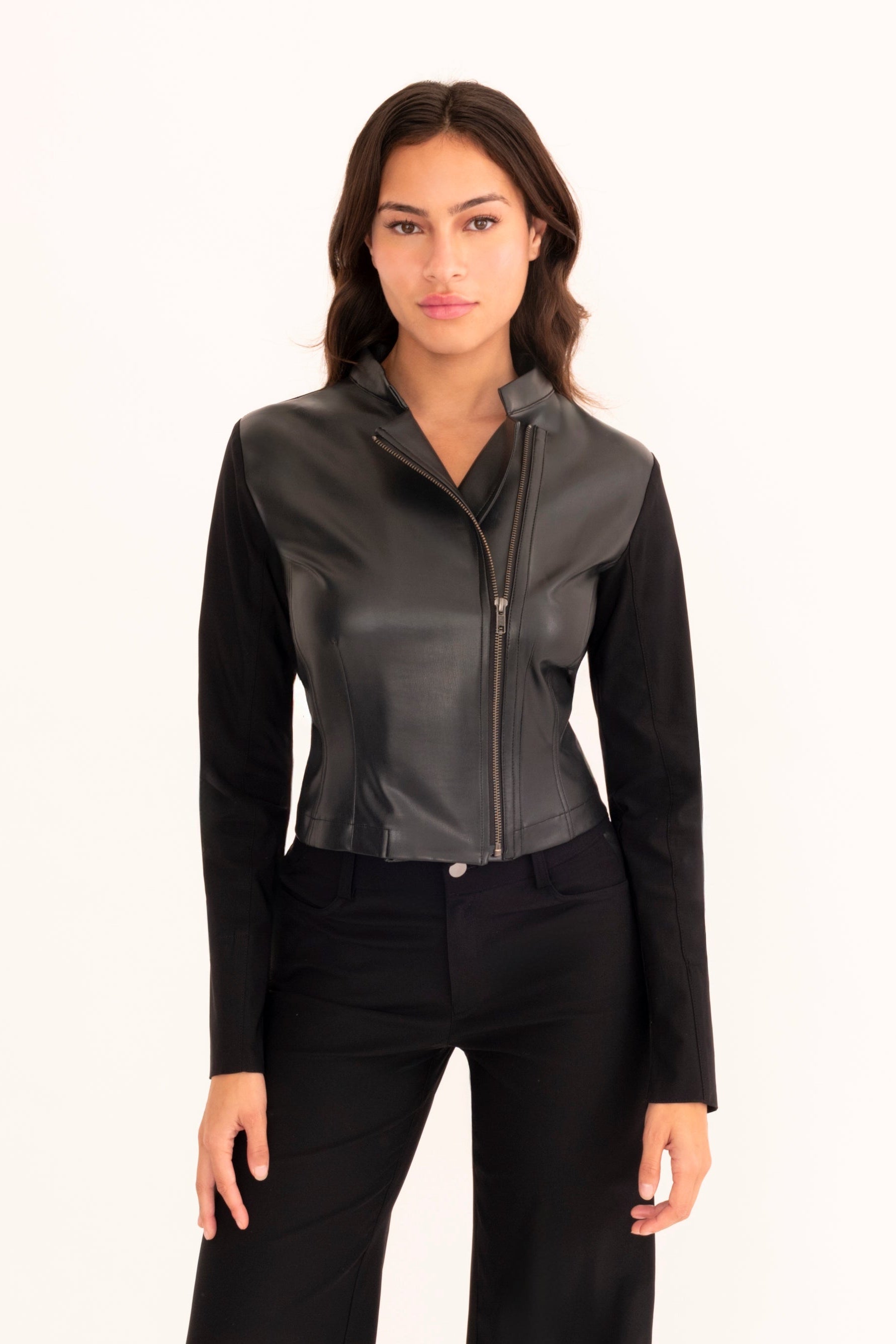 A woman modeling a black faux-leather motorcycle jacket with a stand collar and front zipper, paired with matching black pants, against a plain off-white studio background.