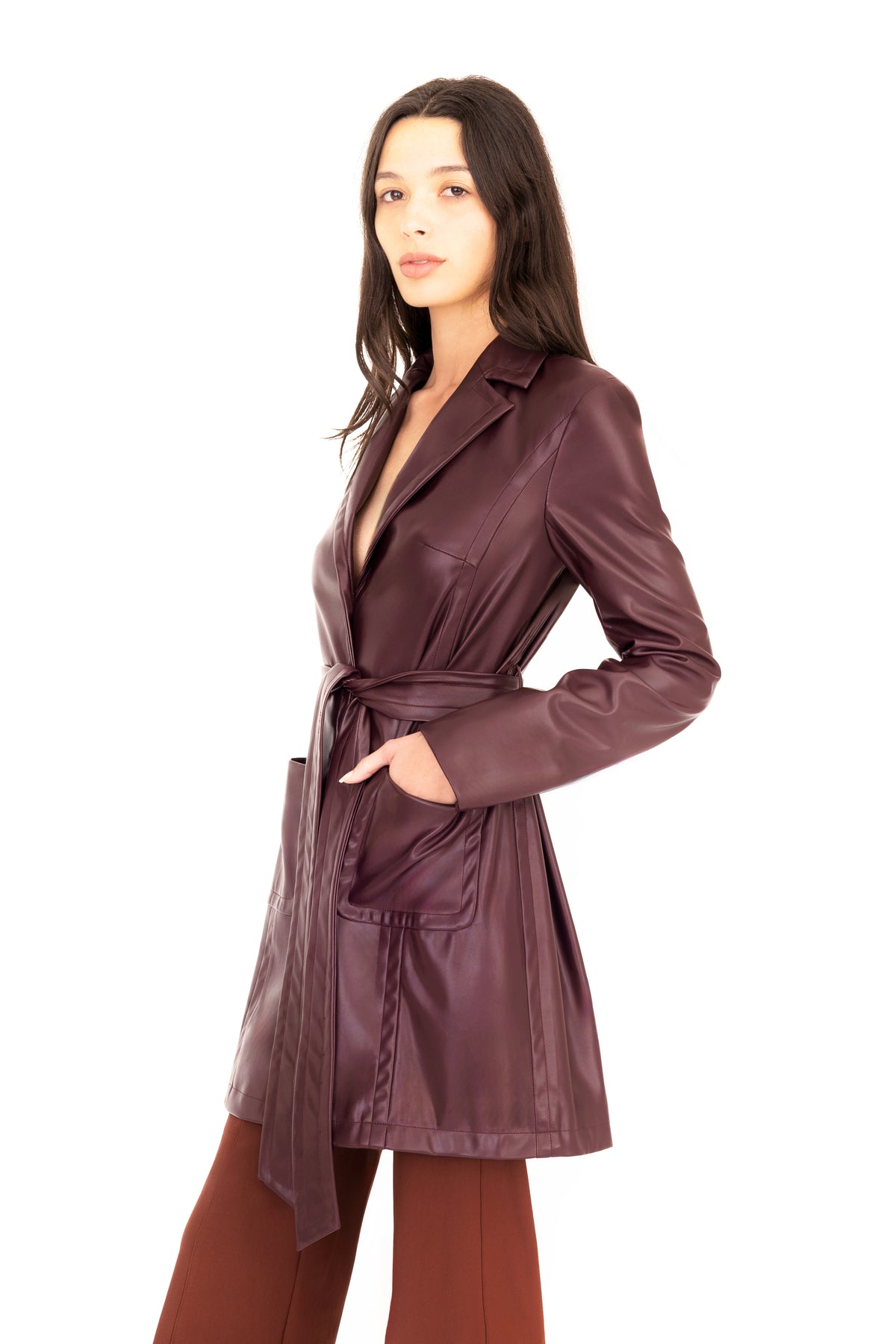 A woman modeling a burgundy vegan leather trench coat with a collar, waist tie, and front pockets, paired with brown pants, against a plain white studio background.