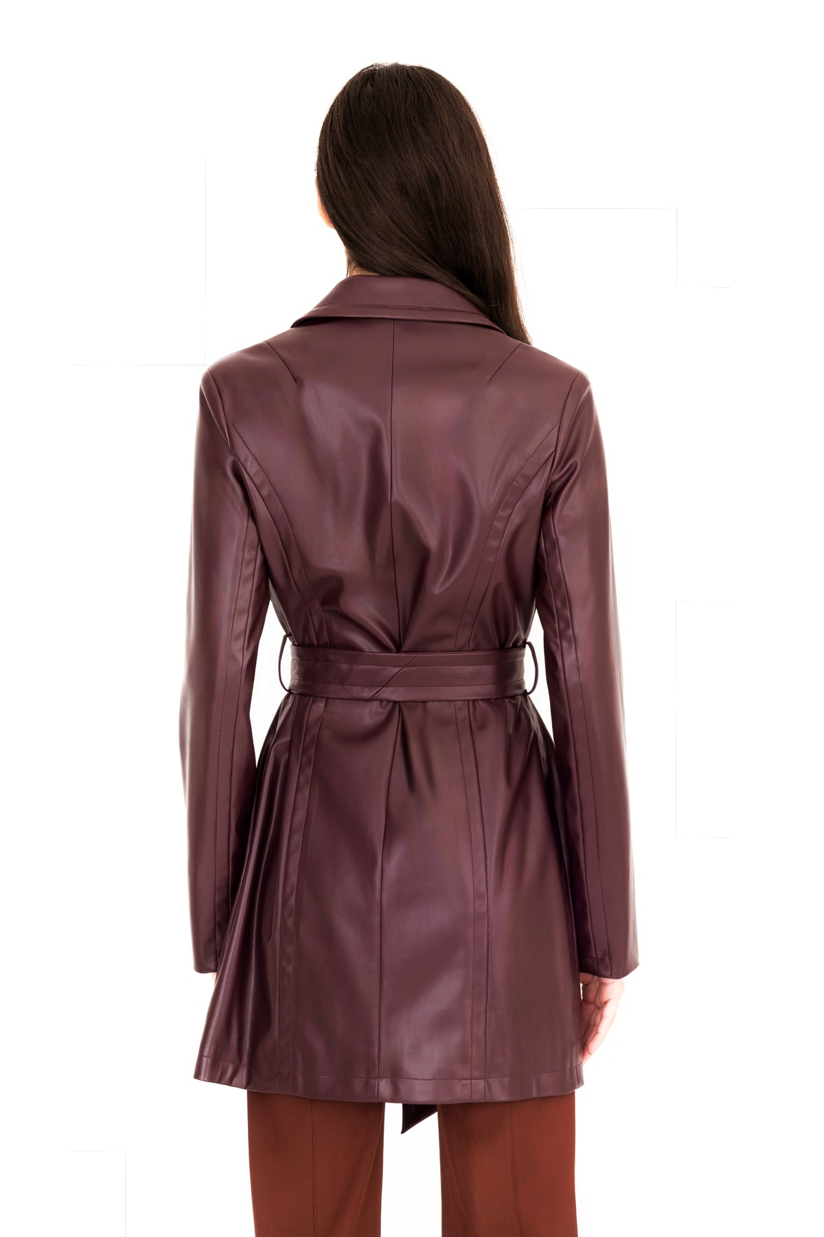 A woman viewed from the back modeling a burgundy vegan leather trench coat with a collar, waist tie, and front pockets, paired with brown pants, against a plain white studio background.