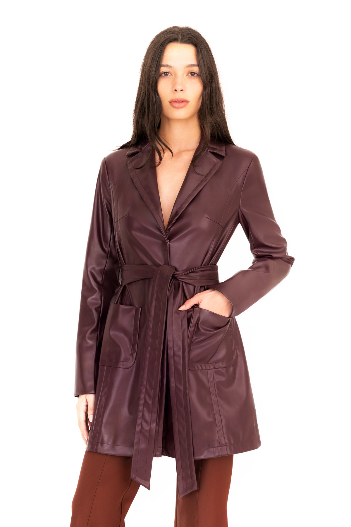 A woman modeling a burgundy vegan leather trench coat with a collar, waist tie, and front pockets, paired with brown pants, against a plain white studio background.