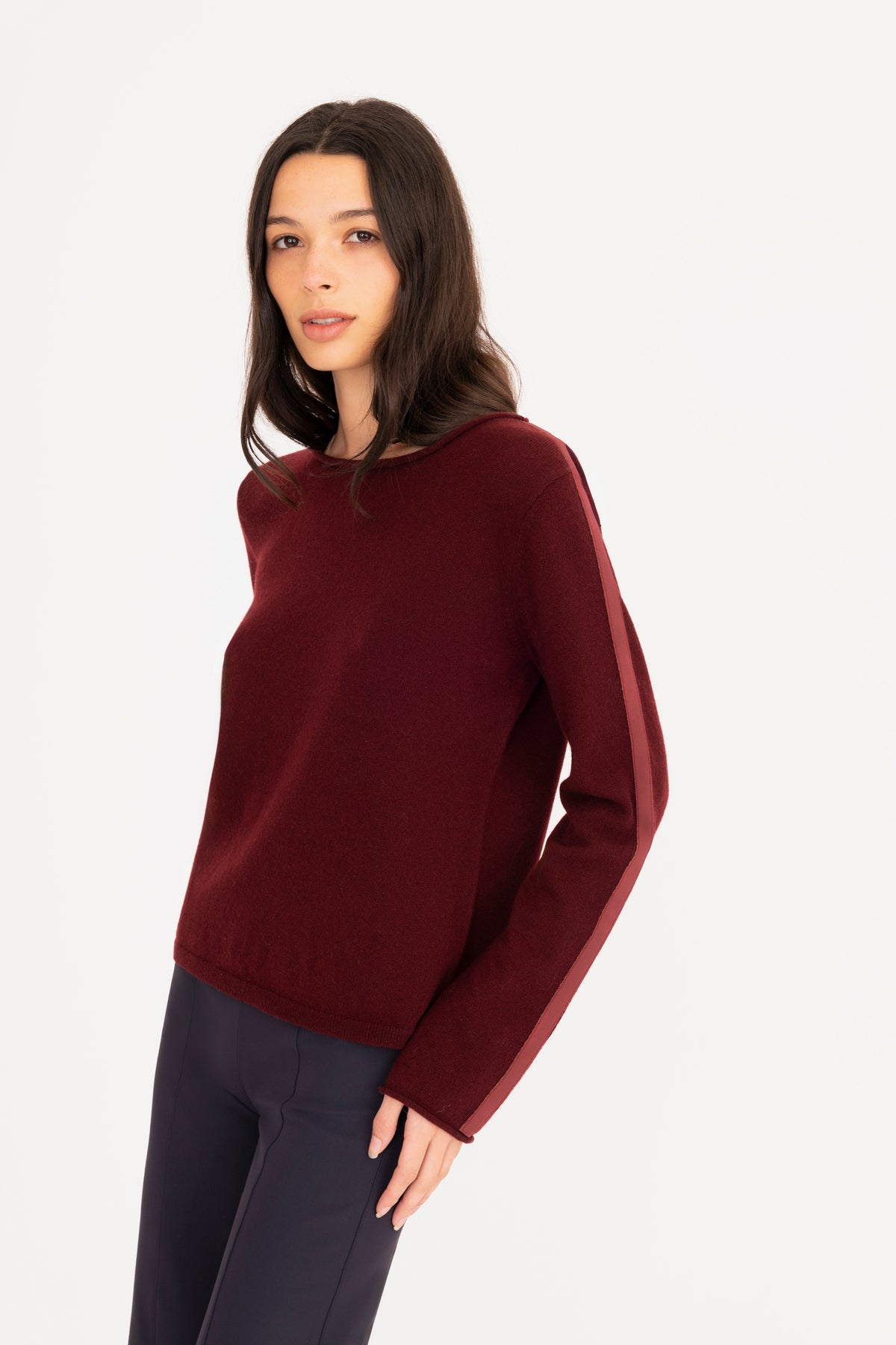 A woman modeling a burgundy long-sleeve knit sweater, paired with navy blue pants, against a plain white studio background.