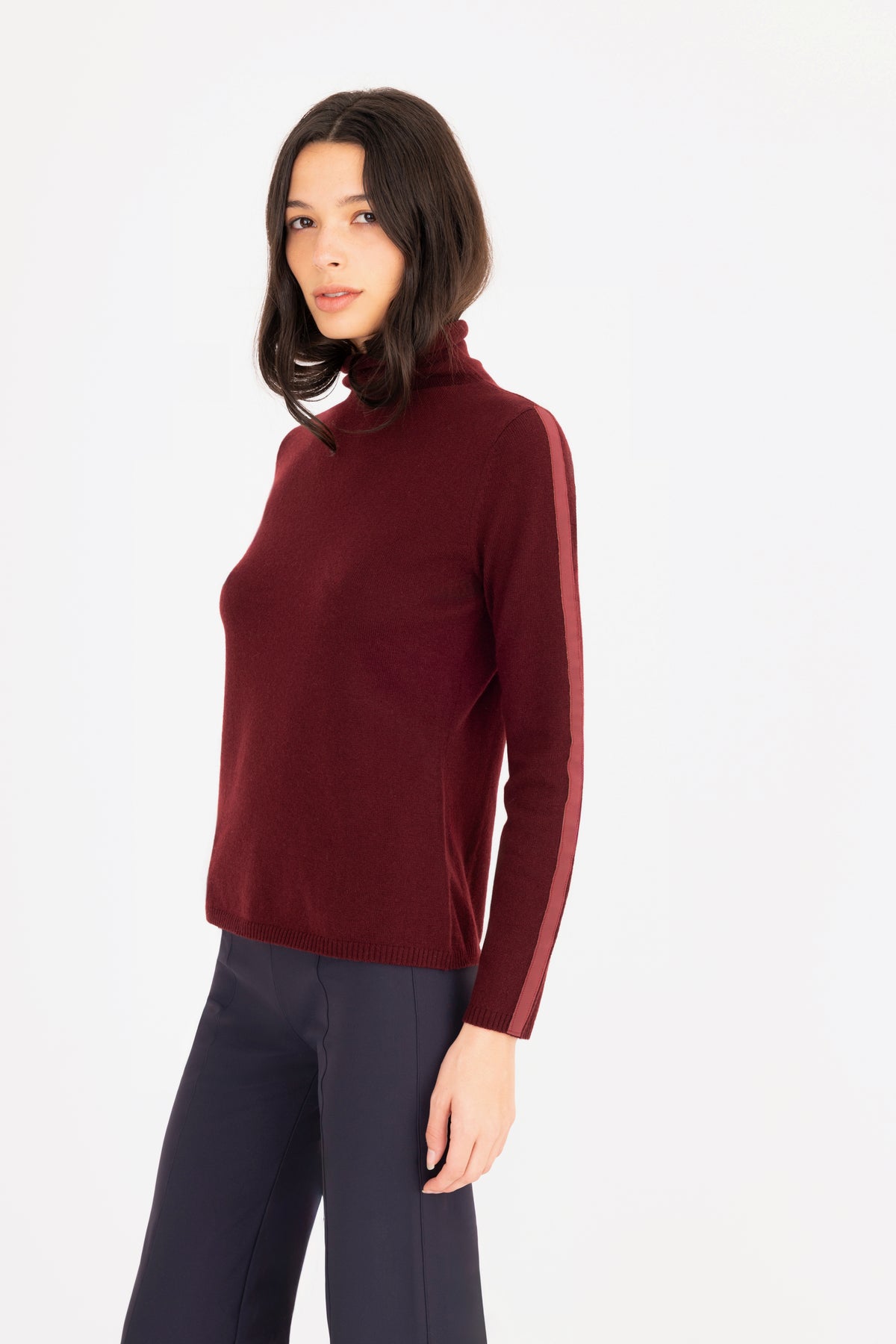 A woman modeling a long-sleeve turtleneck sweater in deep burgundy, paired with dark navy trousers, against a plain white studio background.