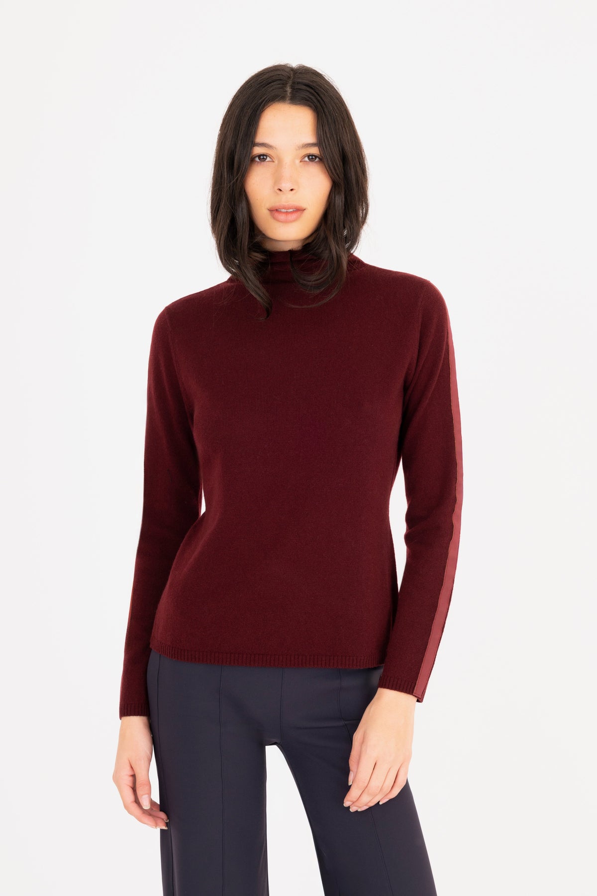 A woman modeling a long-sleeve mock neck sweater in burgundy, paired with dark navy pants, against a plain white studio background.