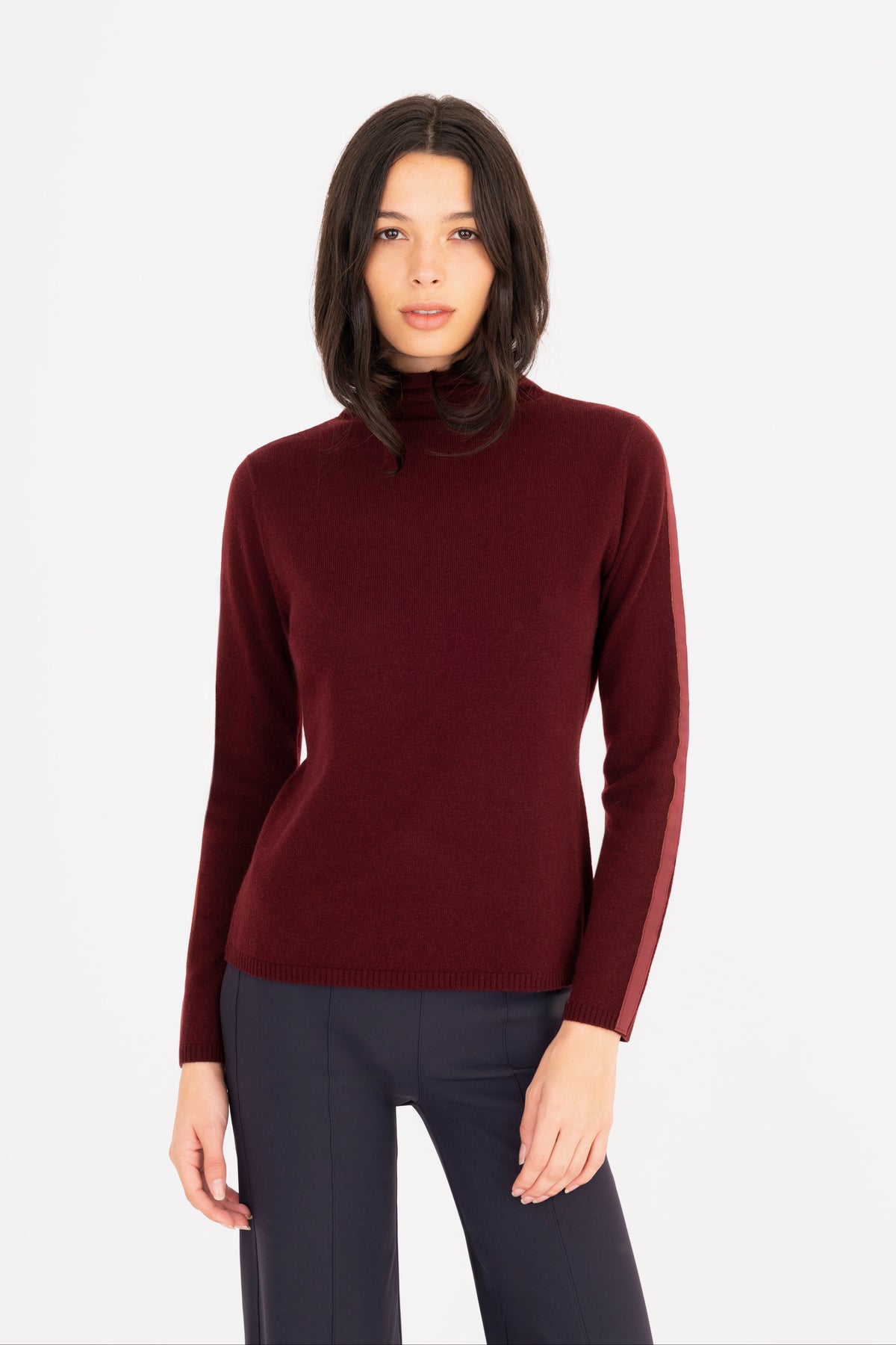 A woman modeling a long-sleeve mock neck sweater in burgundy, paired with dark navy pants, against a plain white studio background.