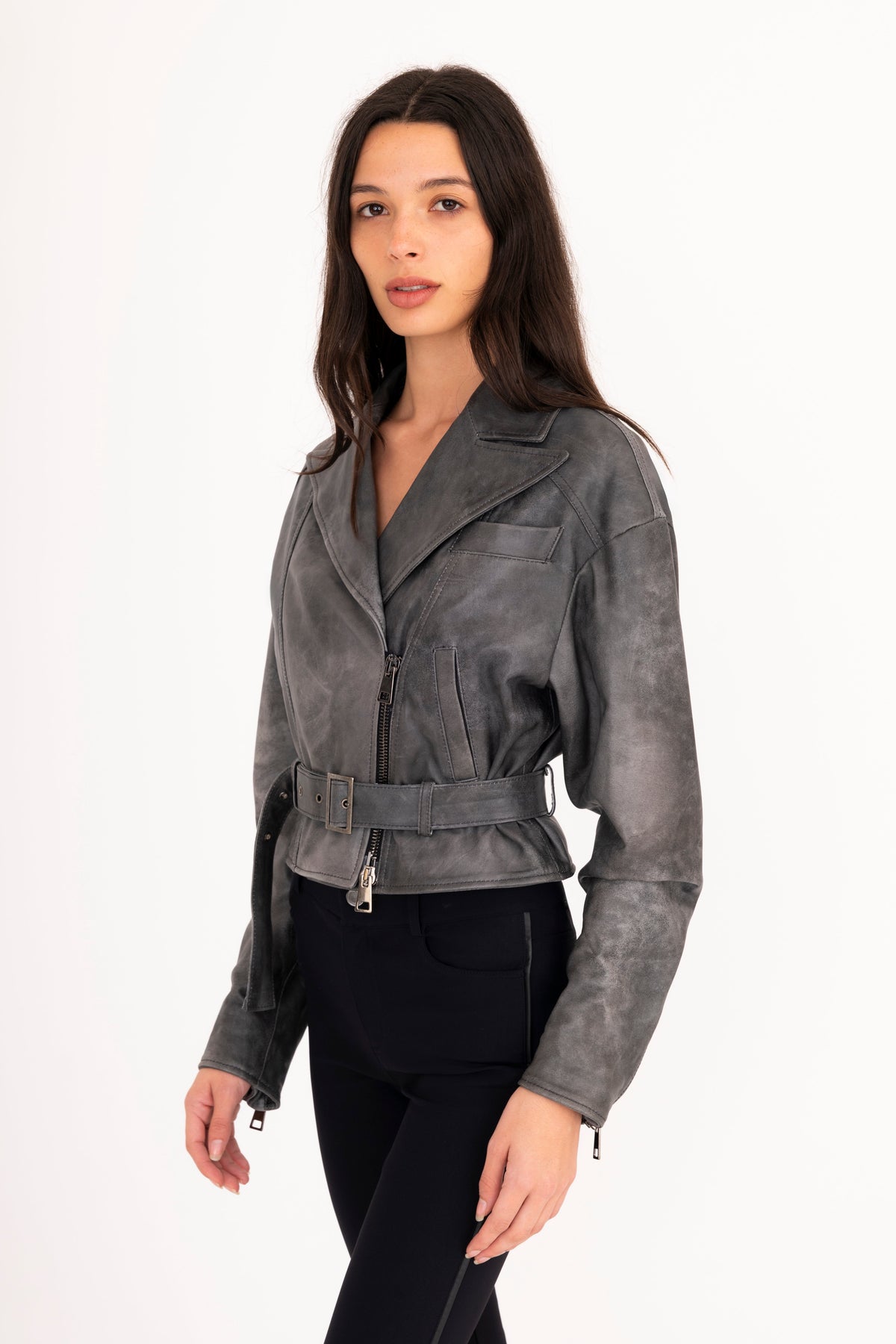 A woman modeling a gray rustic leather moto jacket with a belted waist, paired with black pants, against a plain white studio background.