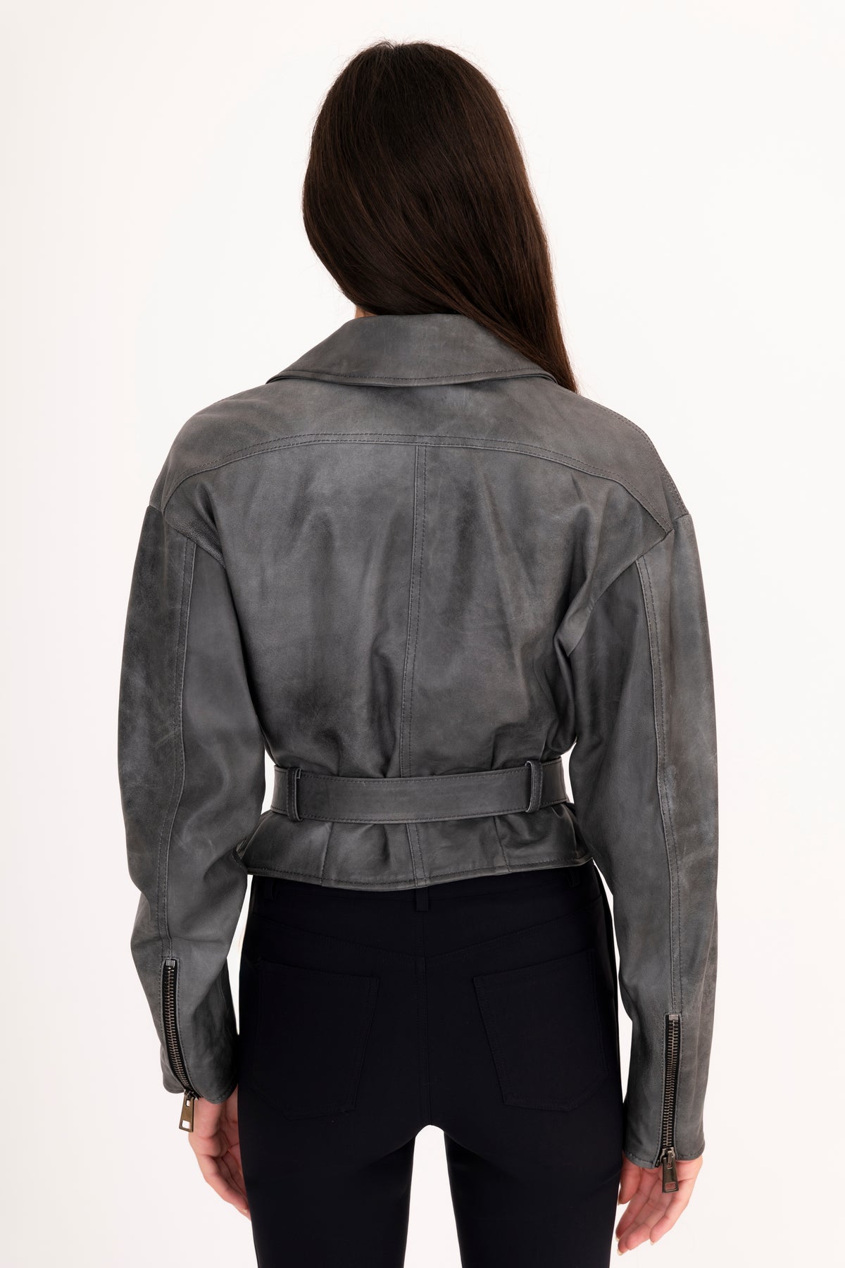 A woman viewed from the back modeling a gray rustic leather moto jacket with a belted waist, paired with black pants, against a plain white studio background.