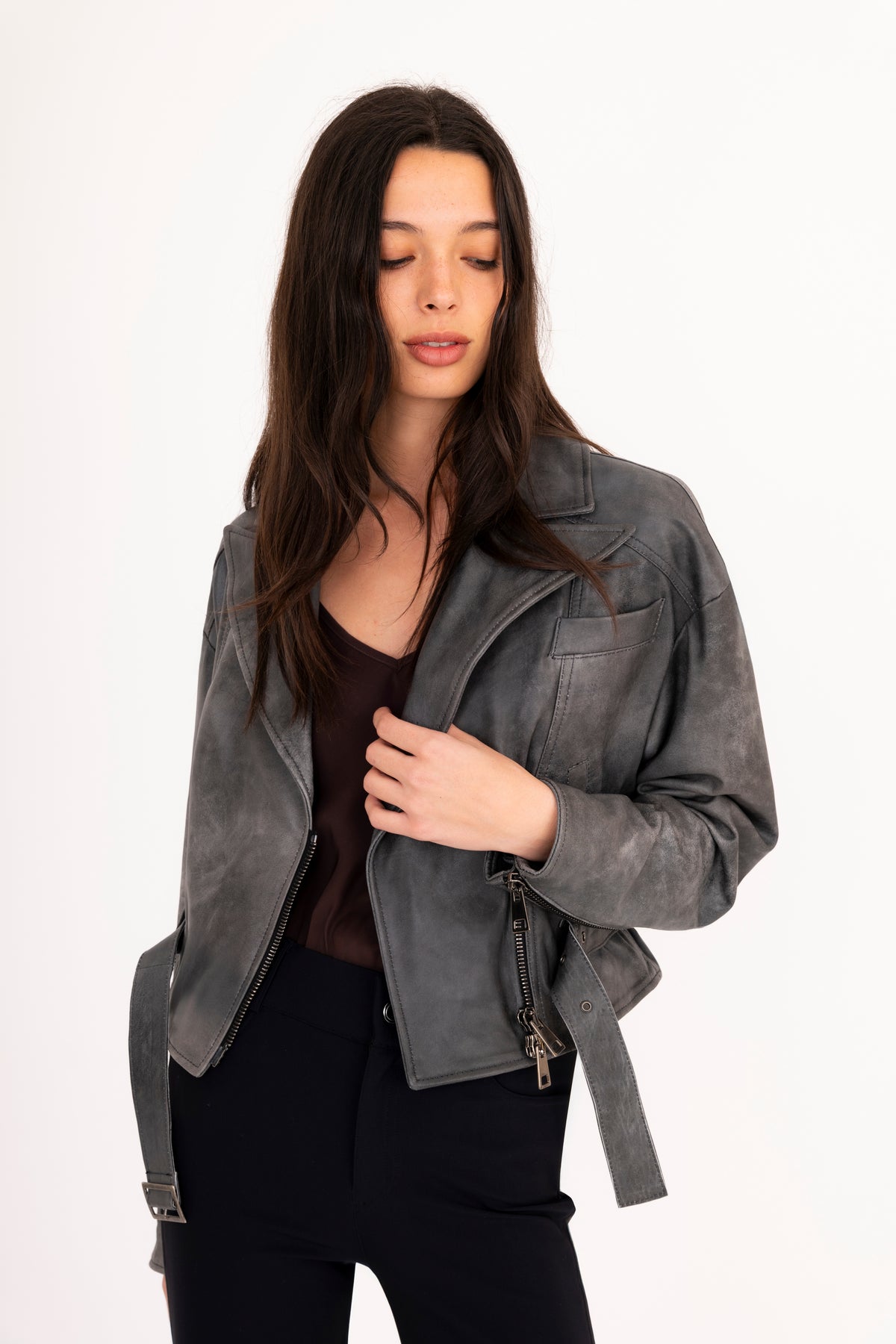 A woman modeling a gray rustic leather moto jacket with a belted waist, paired with black pants, against a plain white studio background.