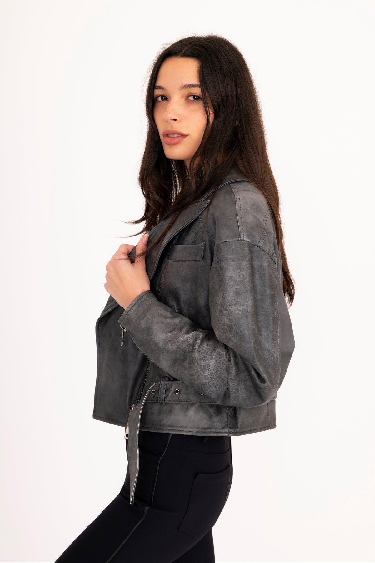 A woman modeling a gray rustic leather moto jacket with a belted waist, paired with black pants, against a plain white studio background.