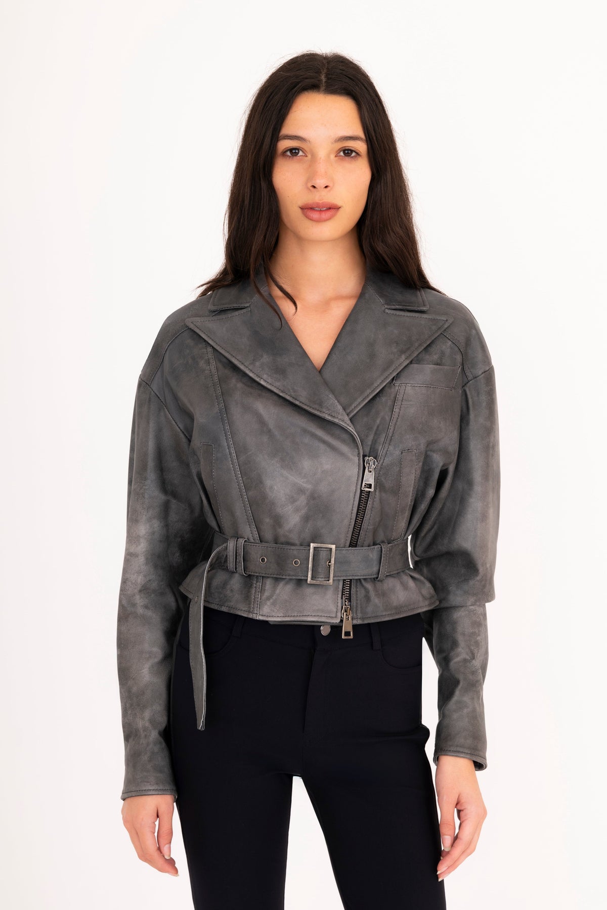 A woman modeling a gray rustic leather moto jacket with a belted waist, paired with black pants, against a plain white studio background.
