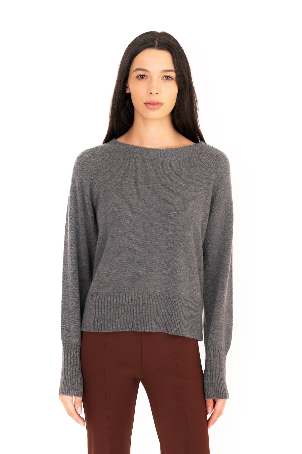 A woman modeling a gray knit crew-neck sweater, paired with dark brown pants, against a plain white background.