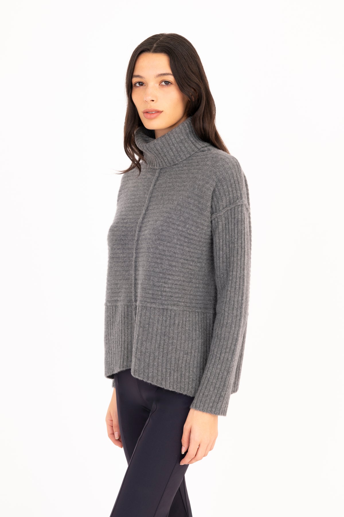 A woman modeling a gray ribbed knit funnel-neck sweater, paired with dark navy pants, against a plain white background.