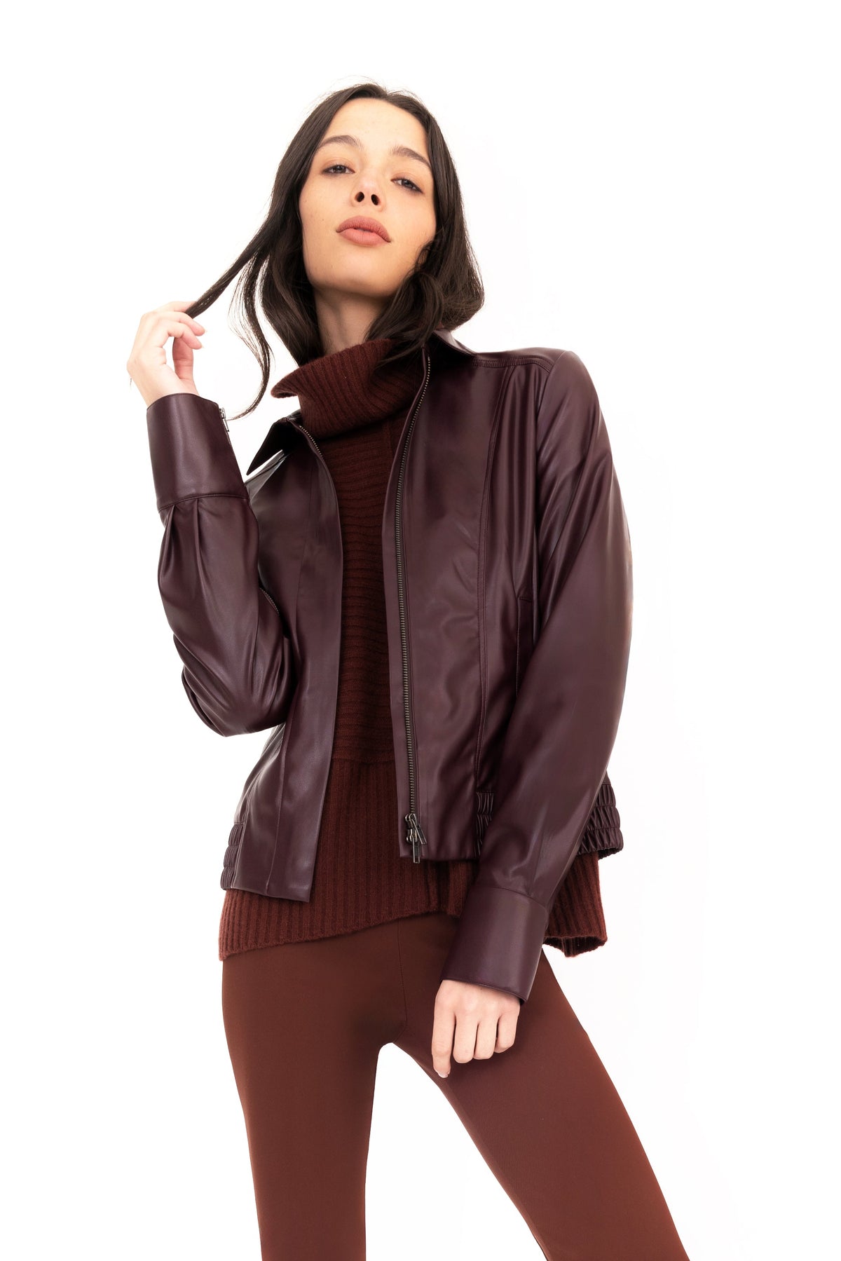 Woman wearing a brown funnel-neck cashmere sweater, burgundy vegan leather jacket, brown pants, and boots on a white background.