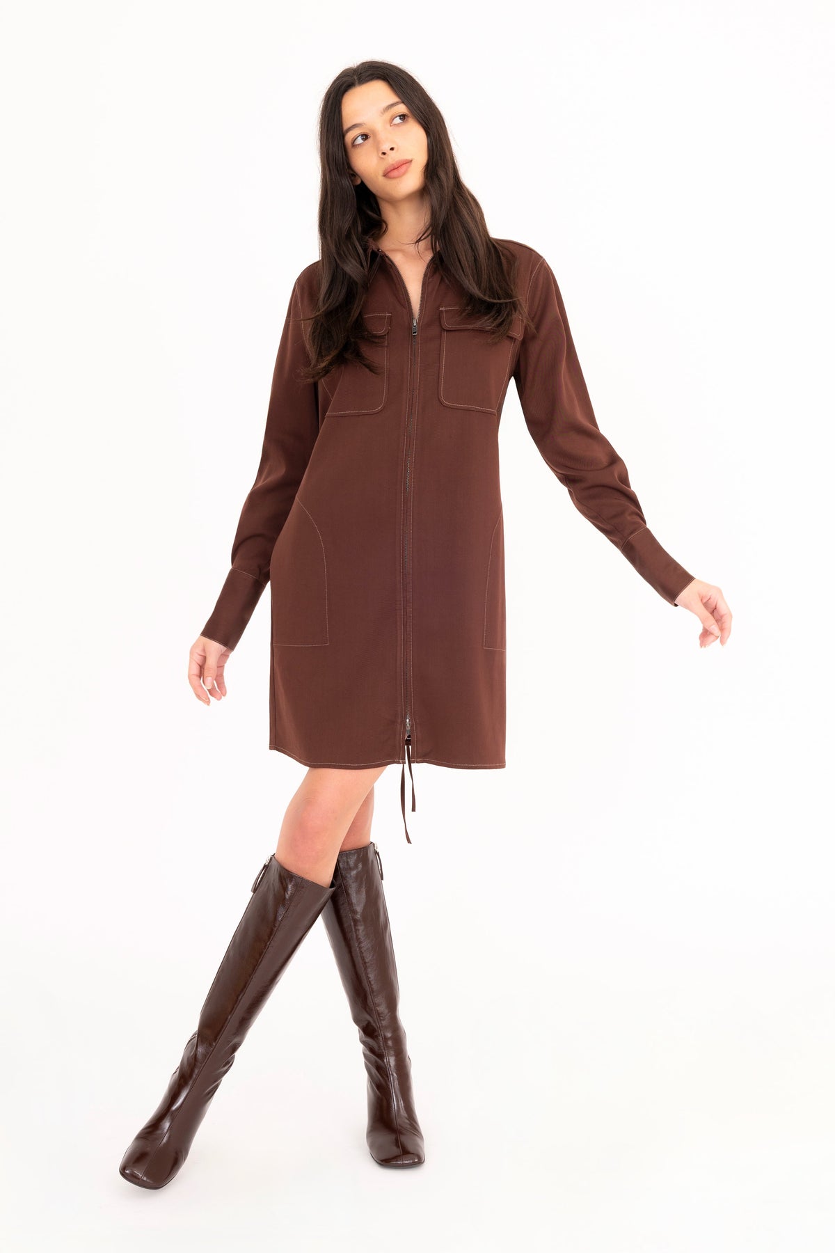 A woman modeling a brown denim shirt dress with front pockets, paired with dark brown knee-high leather boots, against a plain white studio background.