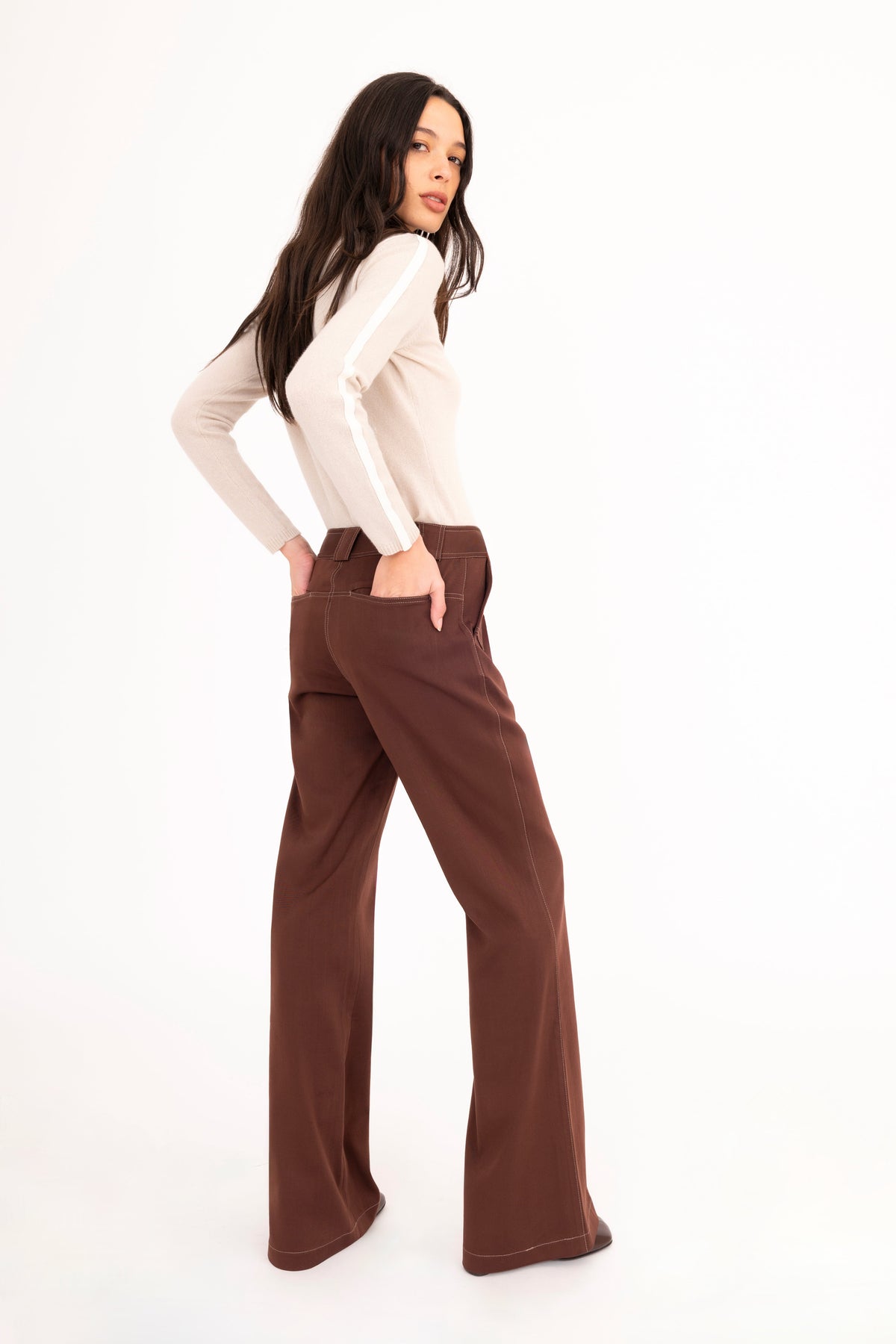 A woman modeling a brown flared denim trousers, and a beige cashmere mock-neck sweater, paired with dark brown leather boots, against a plain white studio background.