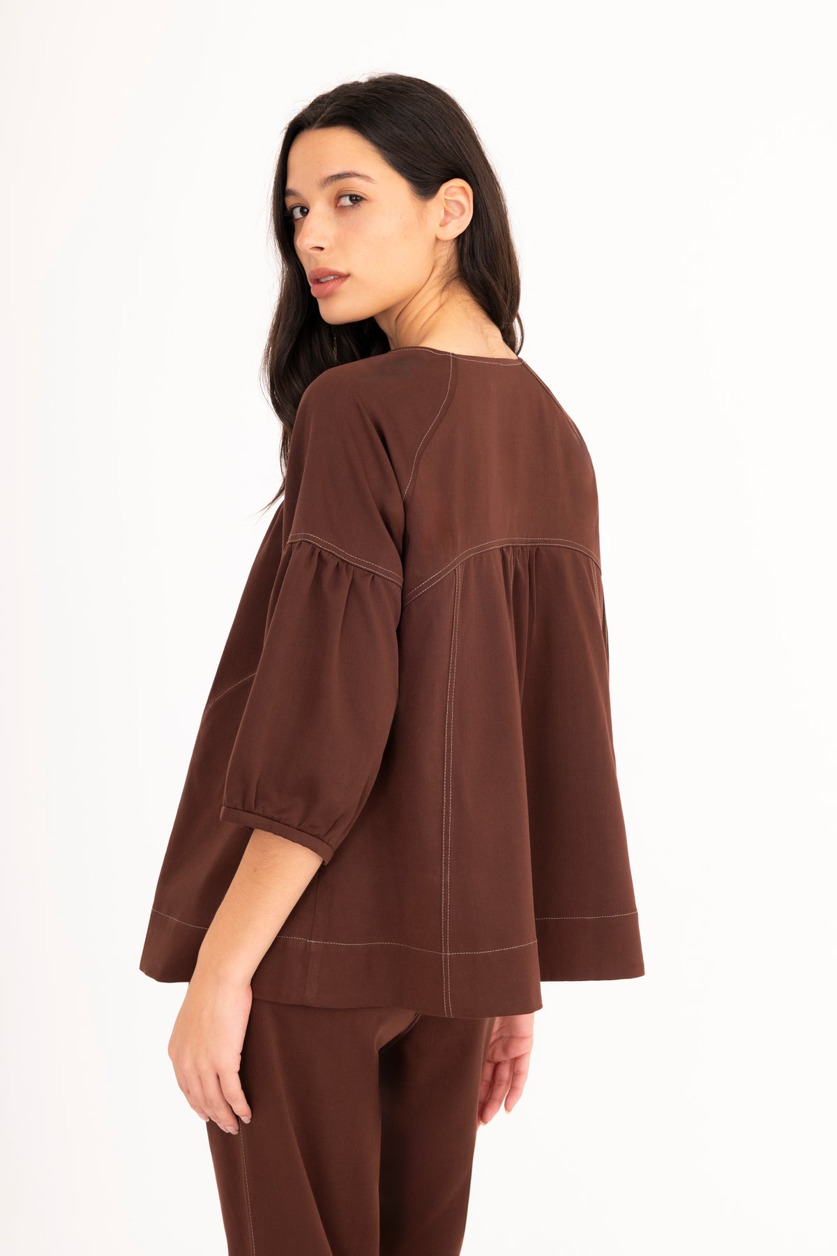 A woman modeling a brown denim top with a split neckline, gathered shoulders, and puff sleeves, paired with matching denim pants against a plain white studio background.