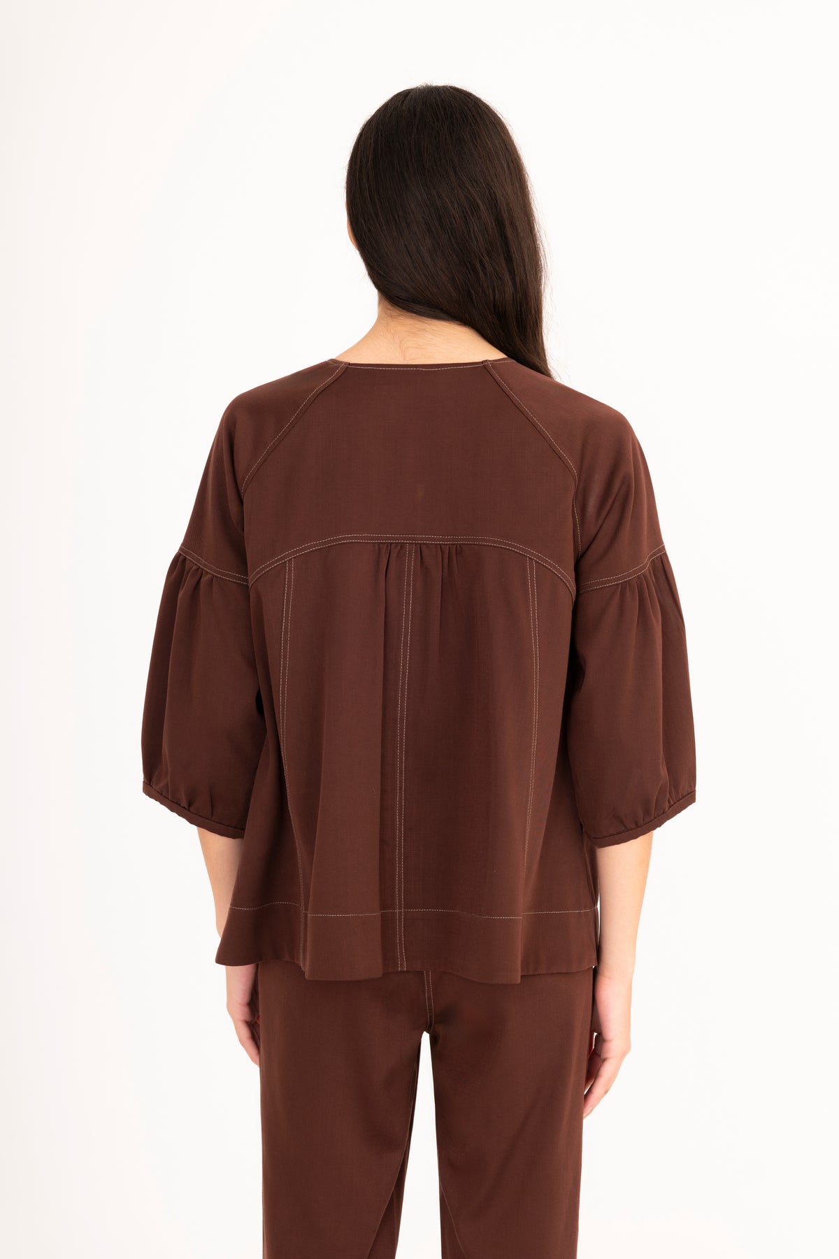 A woman viewed from behind modeling a brown denim top with a split neckline, gathered shoulders, and puff sleeves, paired with matching denim pants against a plain white studio background.