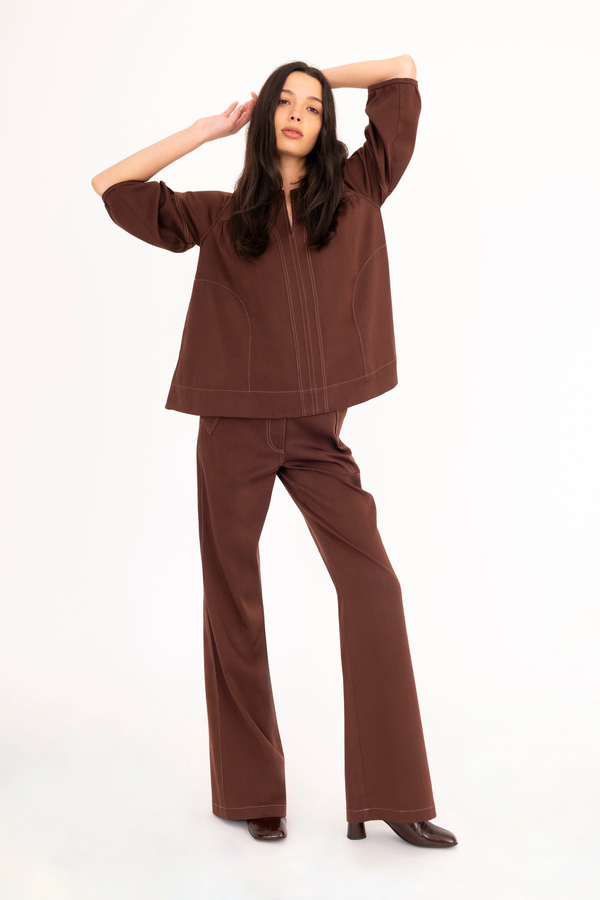 A woman modeling a brown denim top with vertical seam details, paired with brown denim flare pants and brown boots, against a plain white studio background.