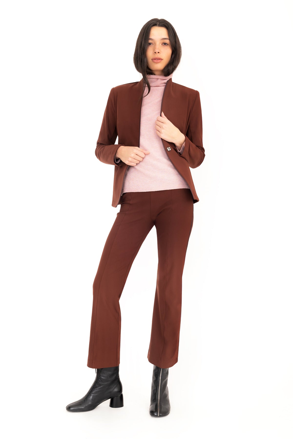 A model poses in a dark brown tailored suit consisting of a blazer and trouser, layered over a light pink turtleneck sweater and paired with black leather ankle boots, against a solid white studio background.