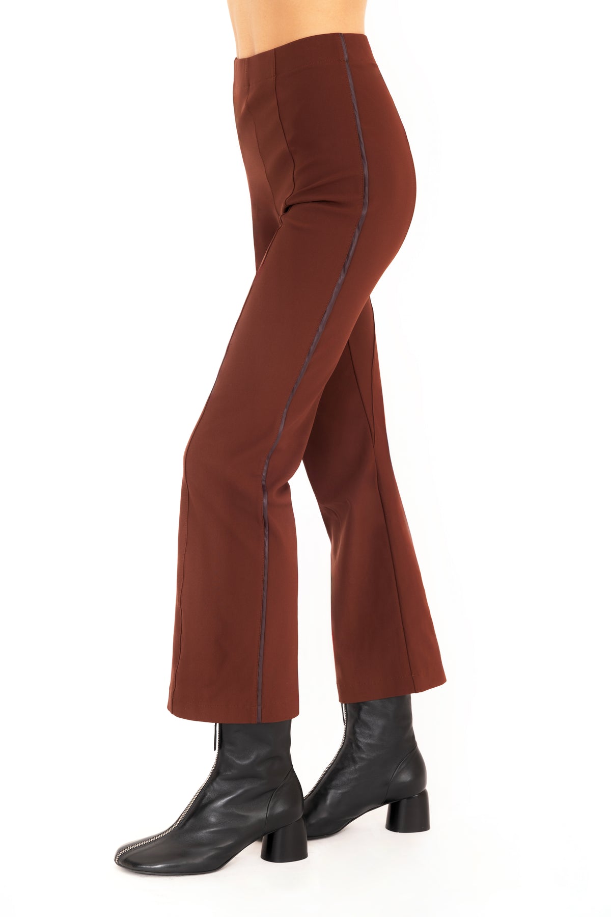 A side profile view of a model wearing dark brown flared pants with dark brown vegan leather contrast piping and black leather ankle boots, set against a plain white background.