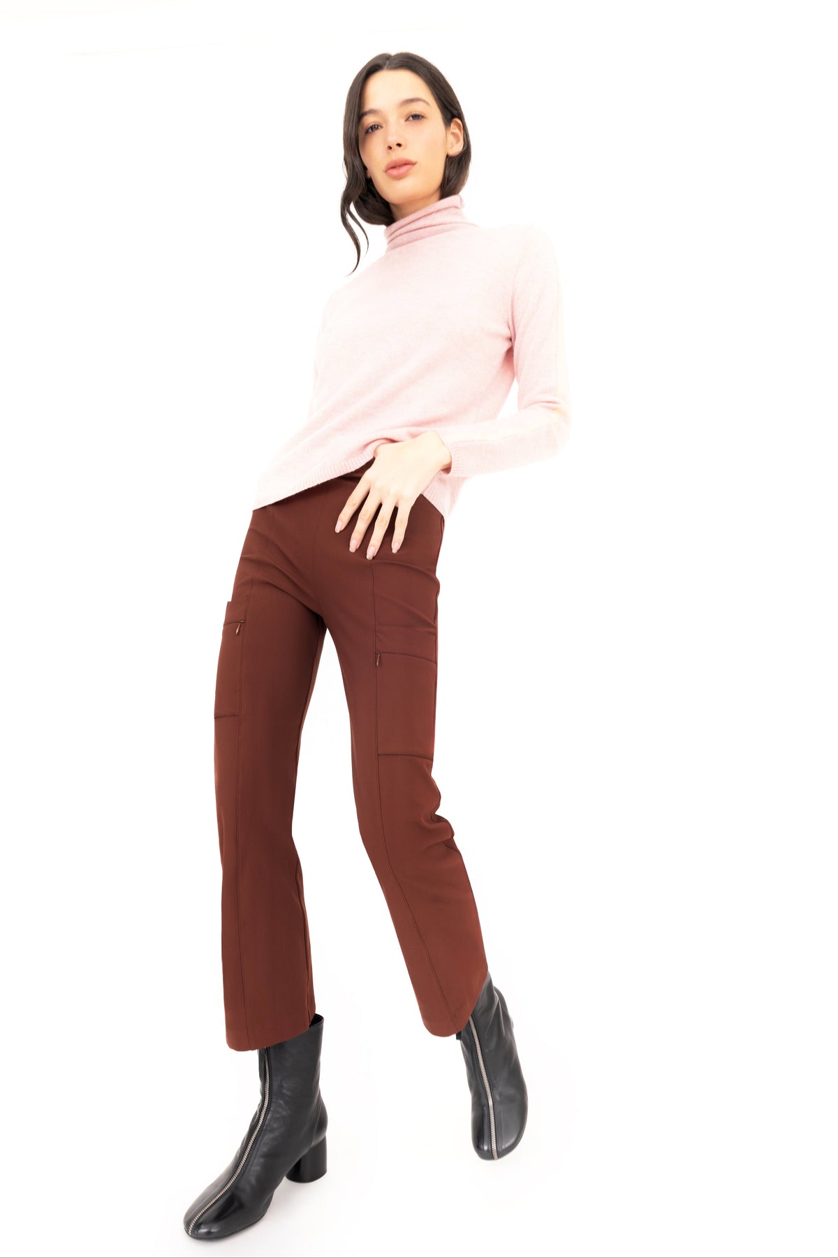 A woman modeling a light pink mock-neck long-sleeve top, brown cropped pants, and black ankle boots against a solid white background.