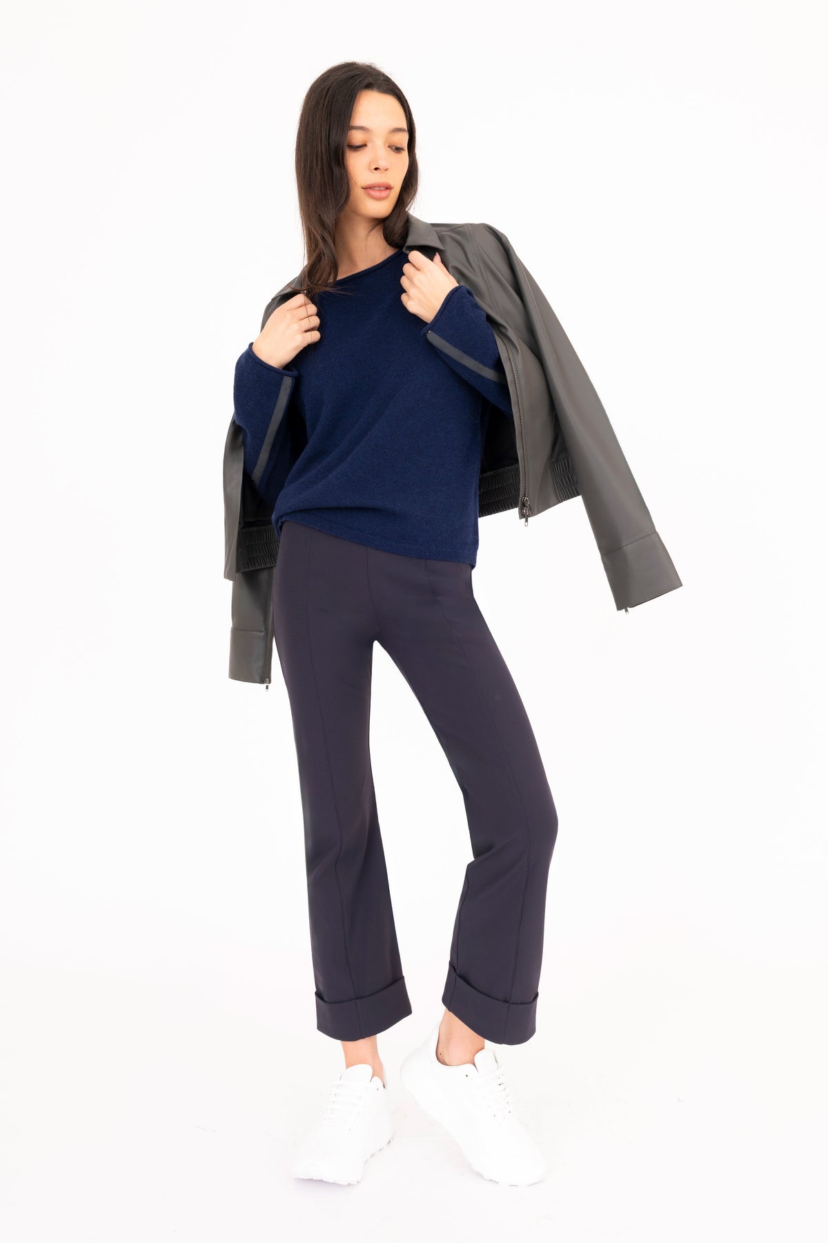 A woman modeling a navy blue cashmere sweater, navy blue cropped pants, white sneakers, and holding a gray vegan leather jacket against her shoulders, shot against a plain white studio background.