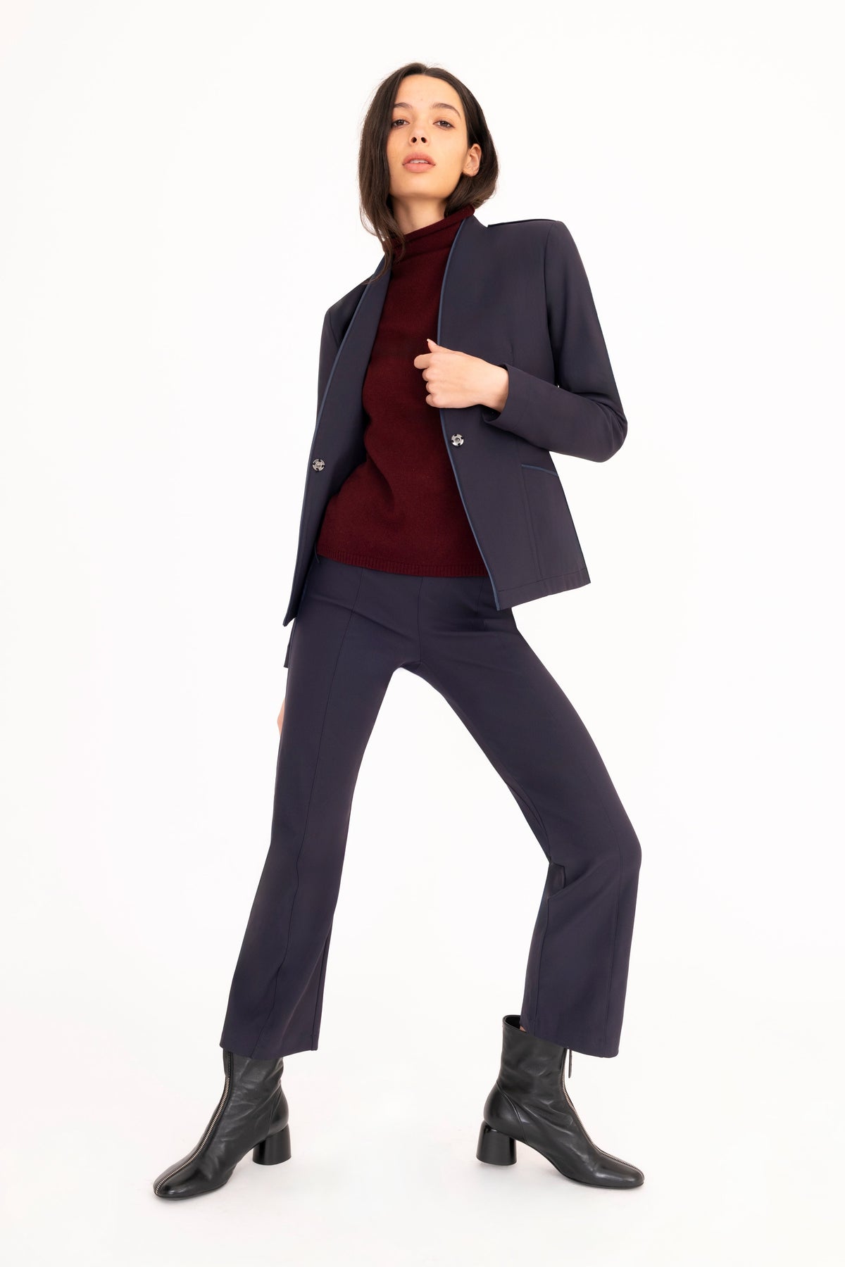 A model poses in a dark navy blue tailored suit consisting of a blazer and trousers, layered over a burgundy turtleneck sweater and paired with black leather ankle boots, against a solid white studio background.