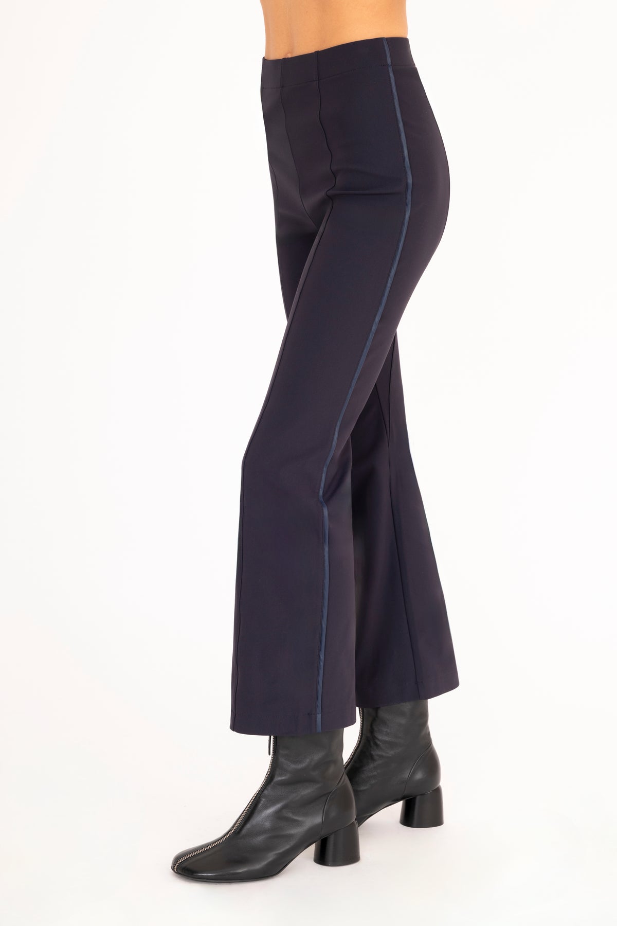 A side view of a model wearing dark blue flared pants with dark blue vegan leather contrast piping and black leather ankle boots, set against a plain white background.