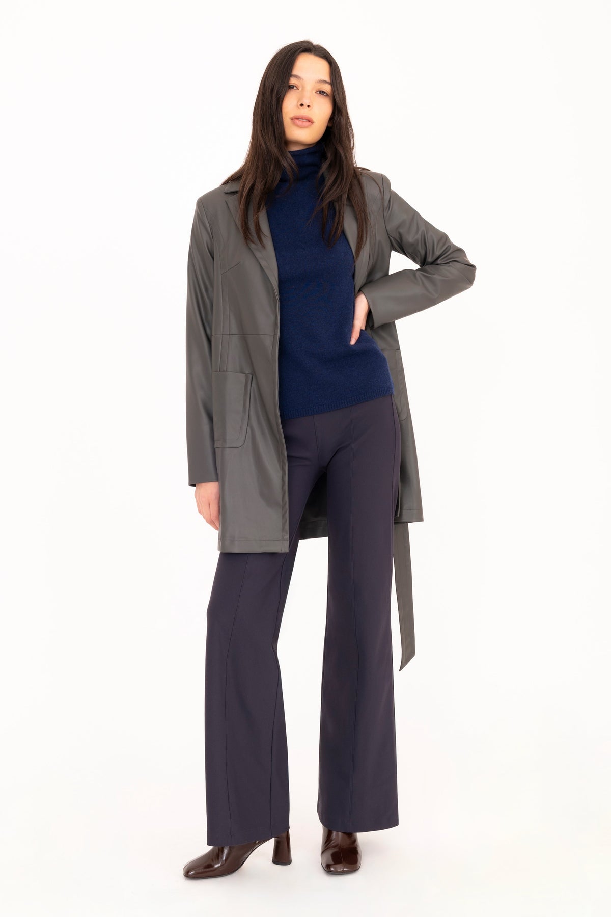 A model wearing a gray vegan leather trench coat over a blue cashmere turtleneck, navy wide-leg trousers, and brown leather boots against a solid white background.