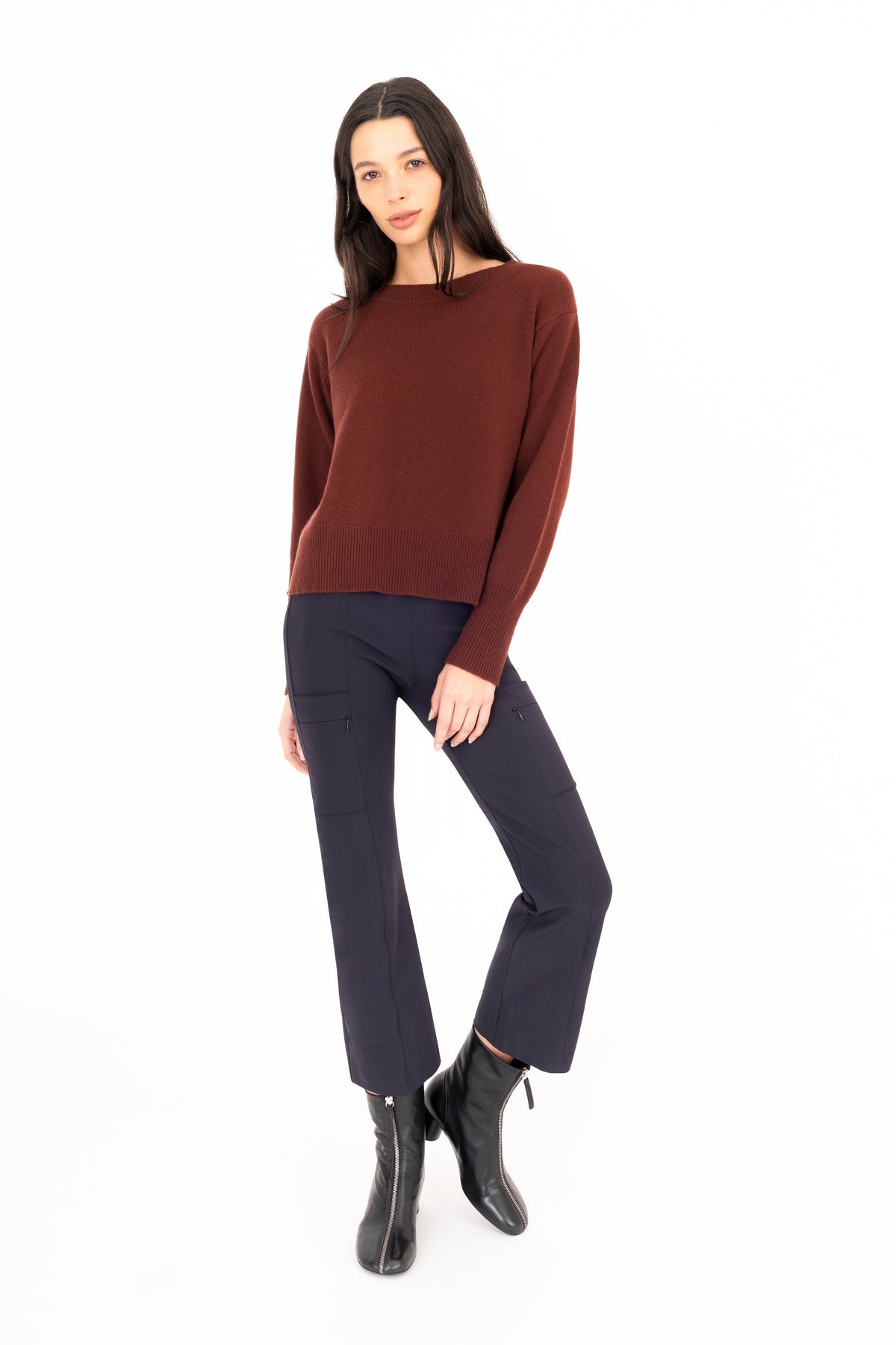 A woman modeling a dark brown cashmere crew-neck sweater, dark navy pants, and black ankle boots against a solid white background.