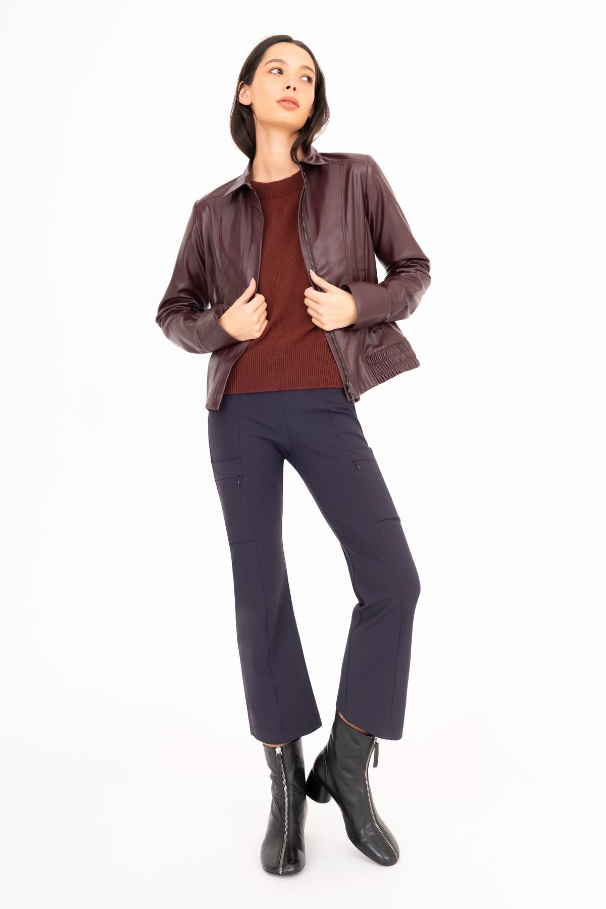 A woman modeling a dark brown leather jacket over a burgundy top, navy blue pants, and black ankle boots, standing against a solid white background.