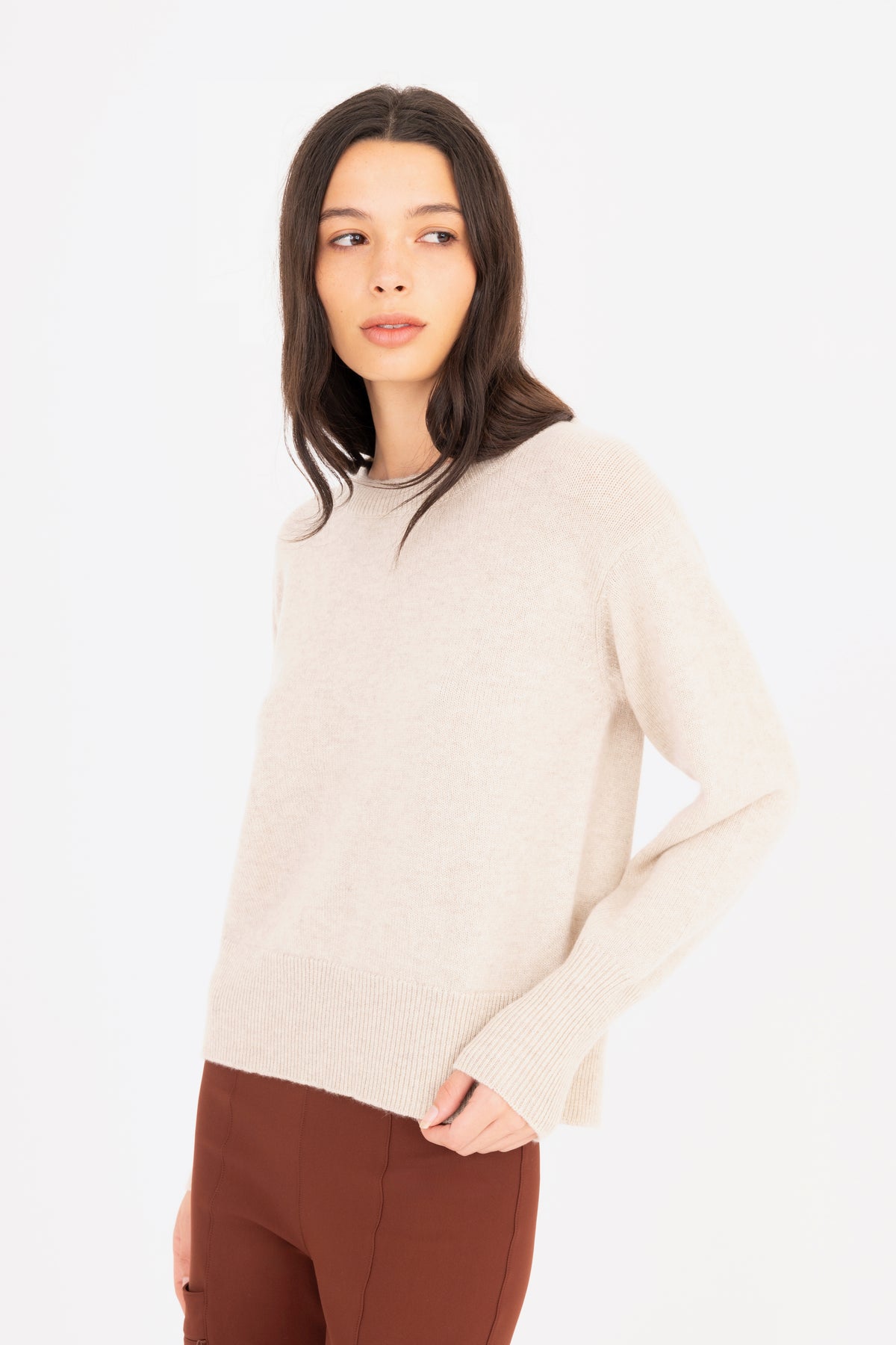 A woman modeling a light beige knit crewneck sweater, paired with brown pants against a plain white studio background.
