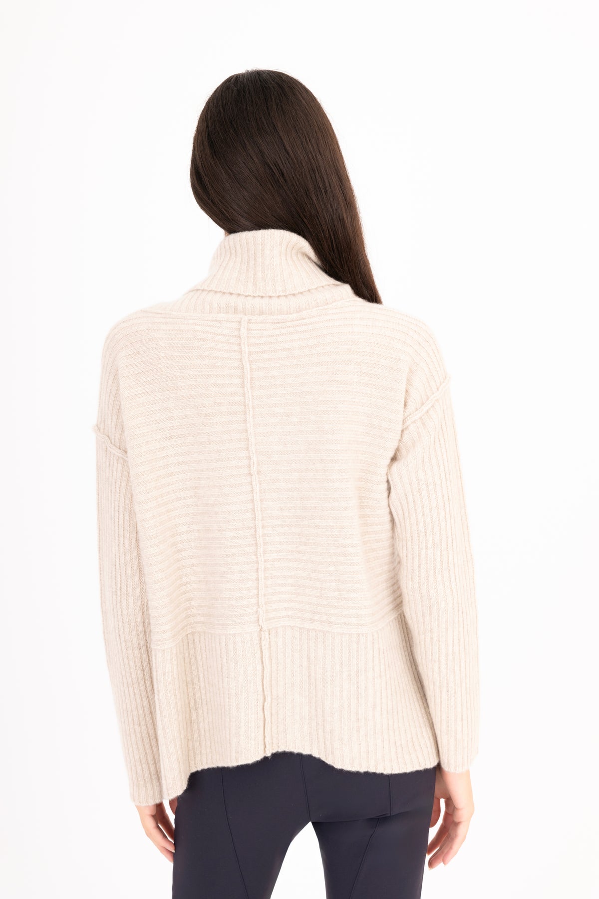 A woman viewed from the back wearing a oatmeal color ribbed knit funnel-neck sweater and dark navy pants against a plain white background.