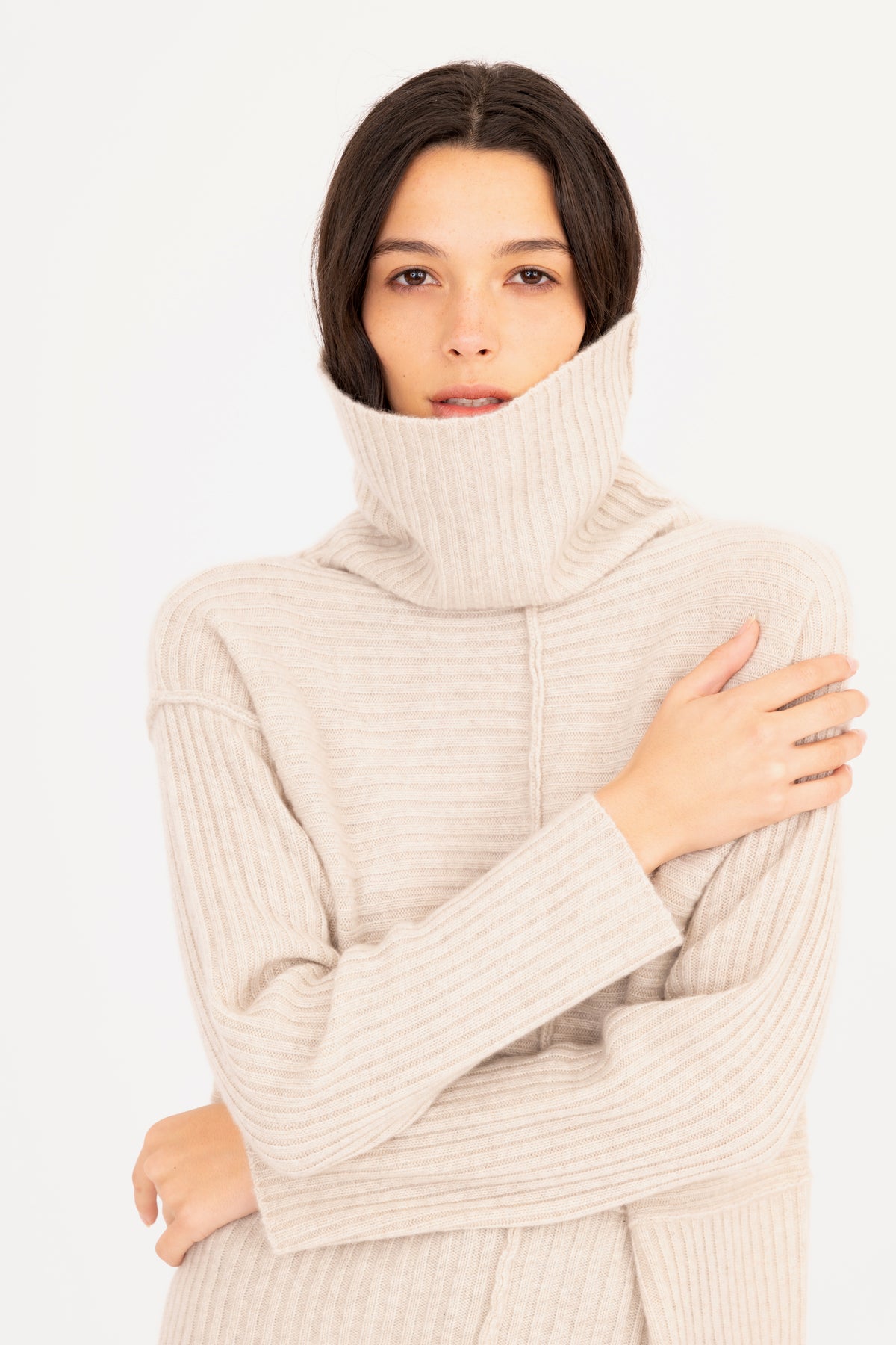 A woman wearing a cream-colored ribbed knit turtleneck sweater, posing with her arms crossed over her chest against a plain white background.