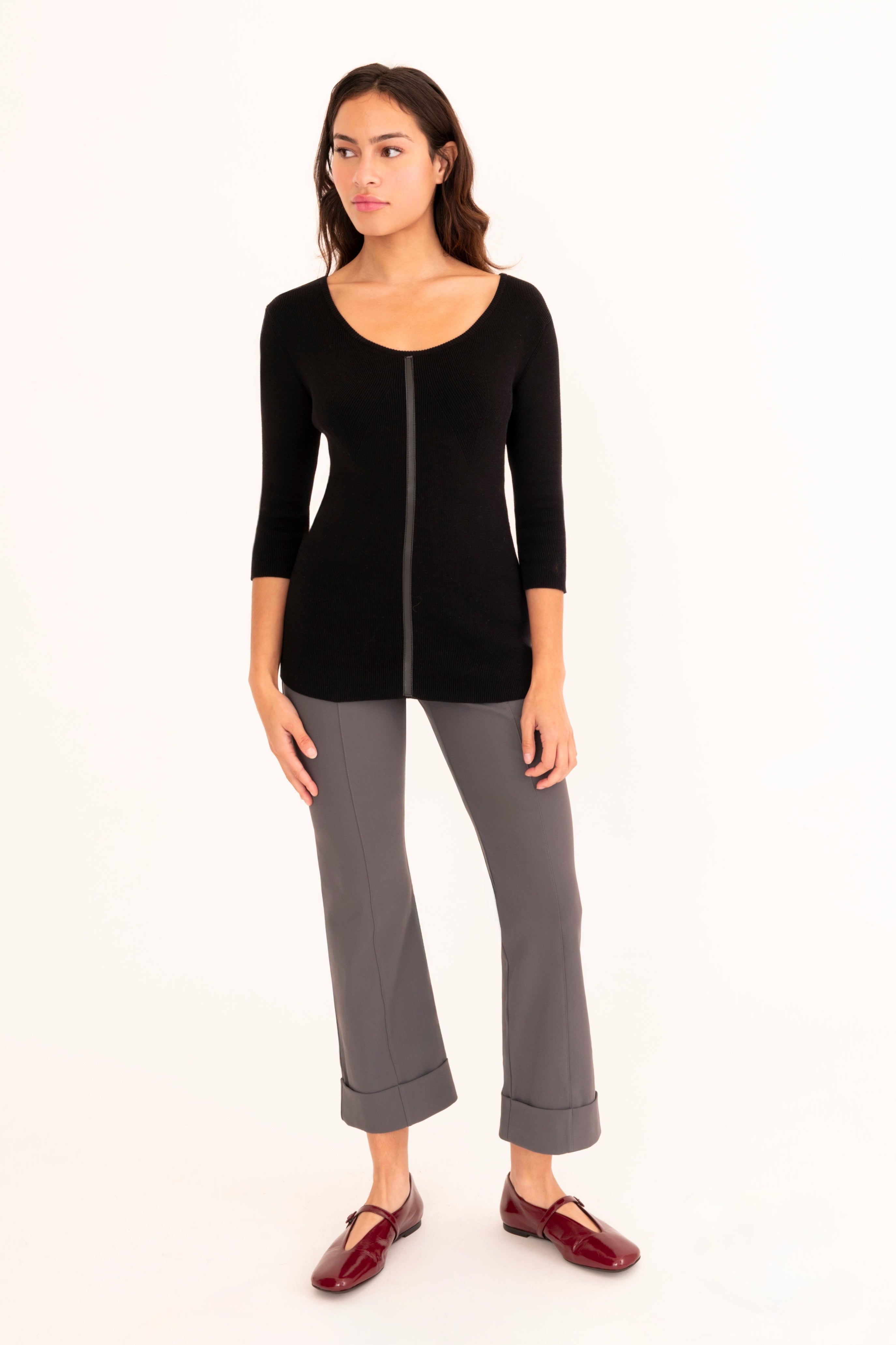 A woman modeling a black three-quarter sleeve knit top paired with gray cropped pants and burgundy loafers, standing against a plain white studio background.