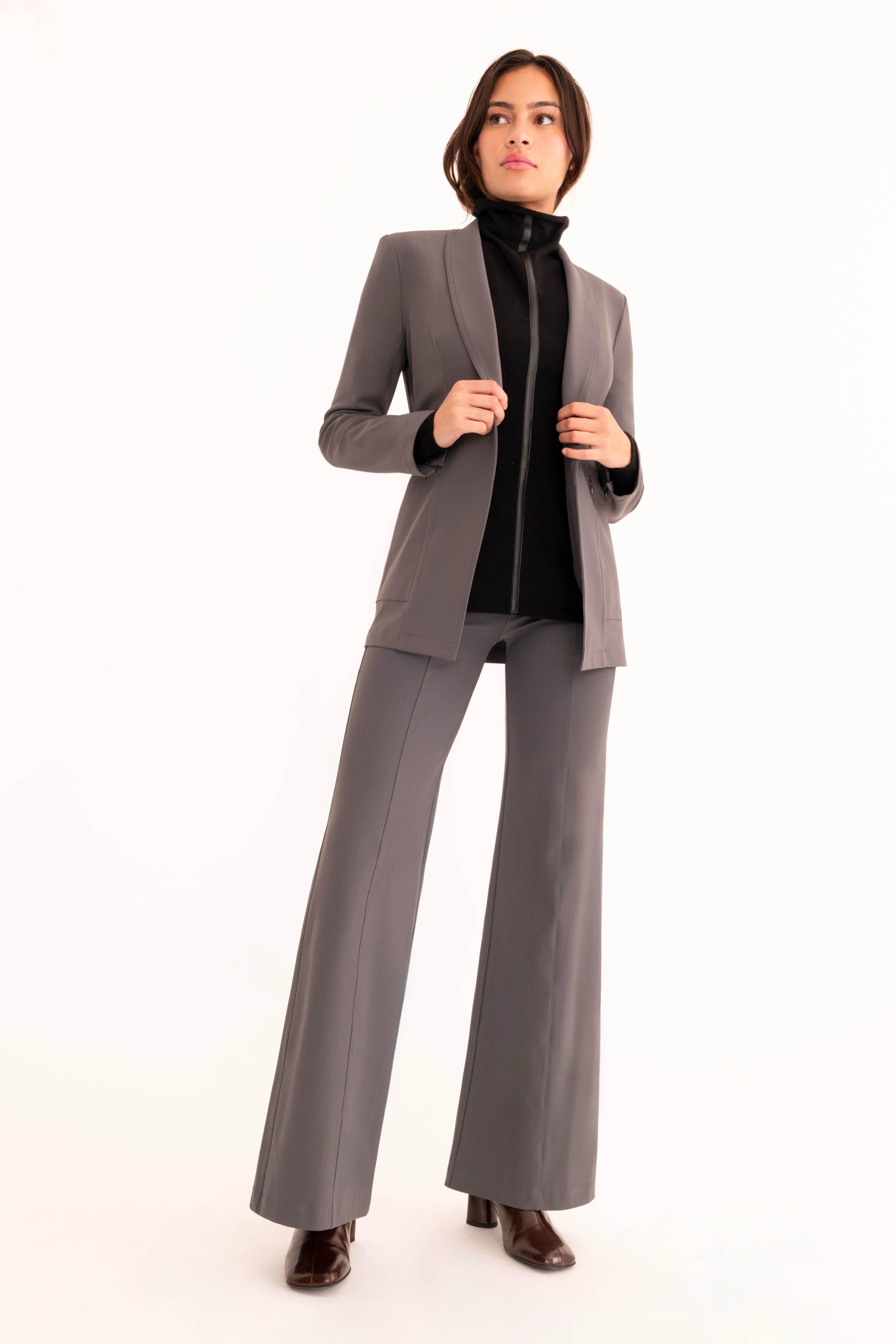 A woman modeling a tailored gray suit with a structured blazer and matching trousers, worn over a gray top and brown ankle boots, photographed against a seamless white studio background.