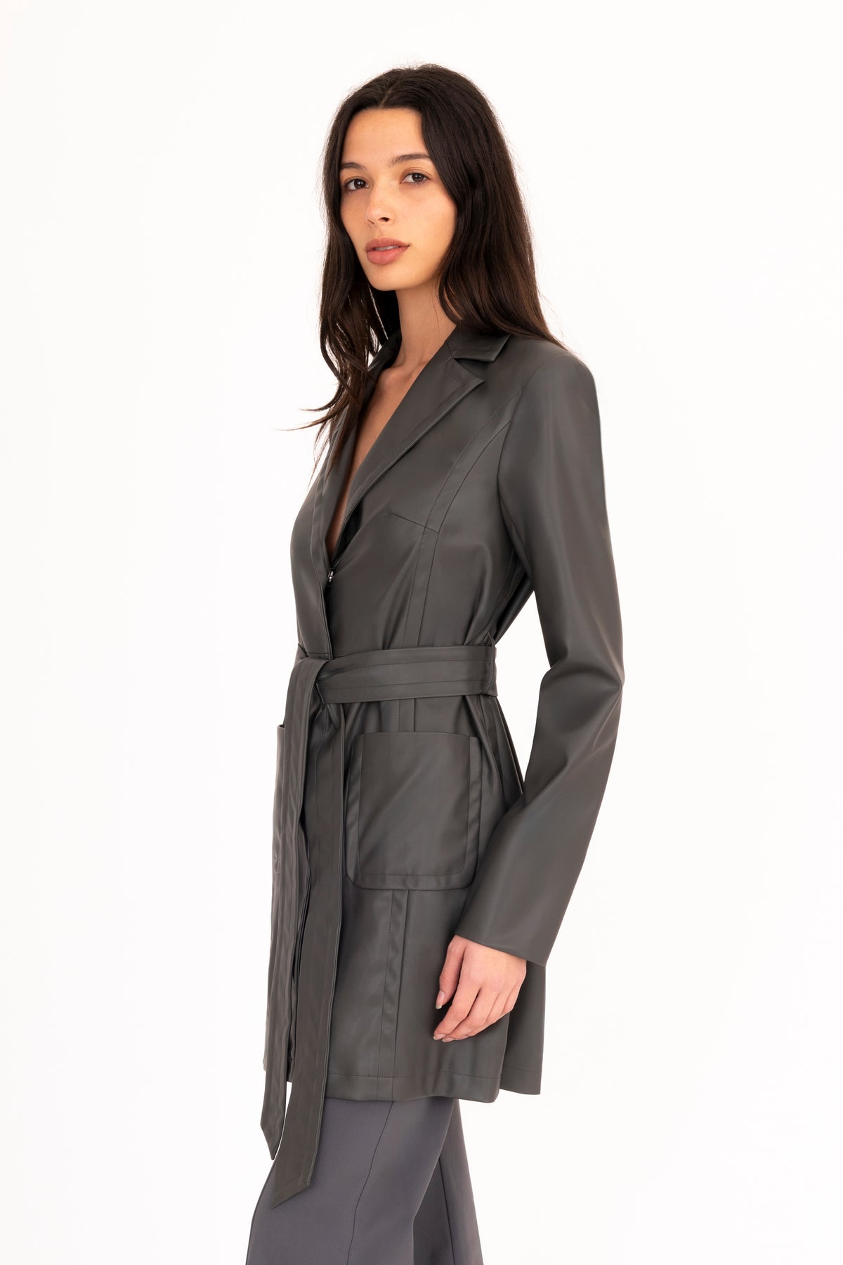 A woman modeling a dark gray vegan leather trench coat with a collar, waist tie, and front pockets, paired with matching dark gray pants, against a plain white studio background.