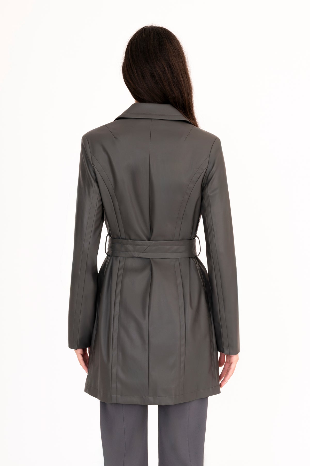 A woman viewed from the back modeling a dark gray vegan leather trench coat with a collar, waist tie, and front pockets, paired with matching dark gray pants, against a plain white studio background.