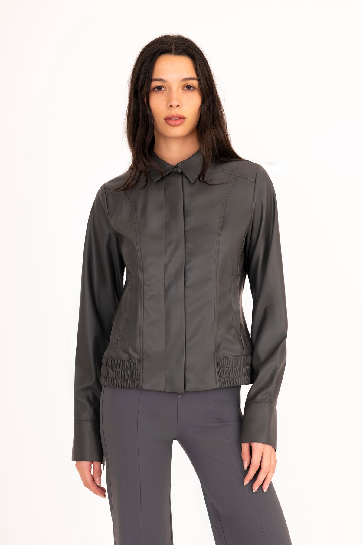 A woman modeling a dark gray vegan leather jacket with a collar, front zipper, and side pockets, paired with matching gray pants, against a plain white studio background.