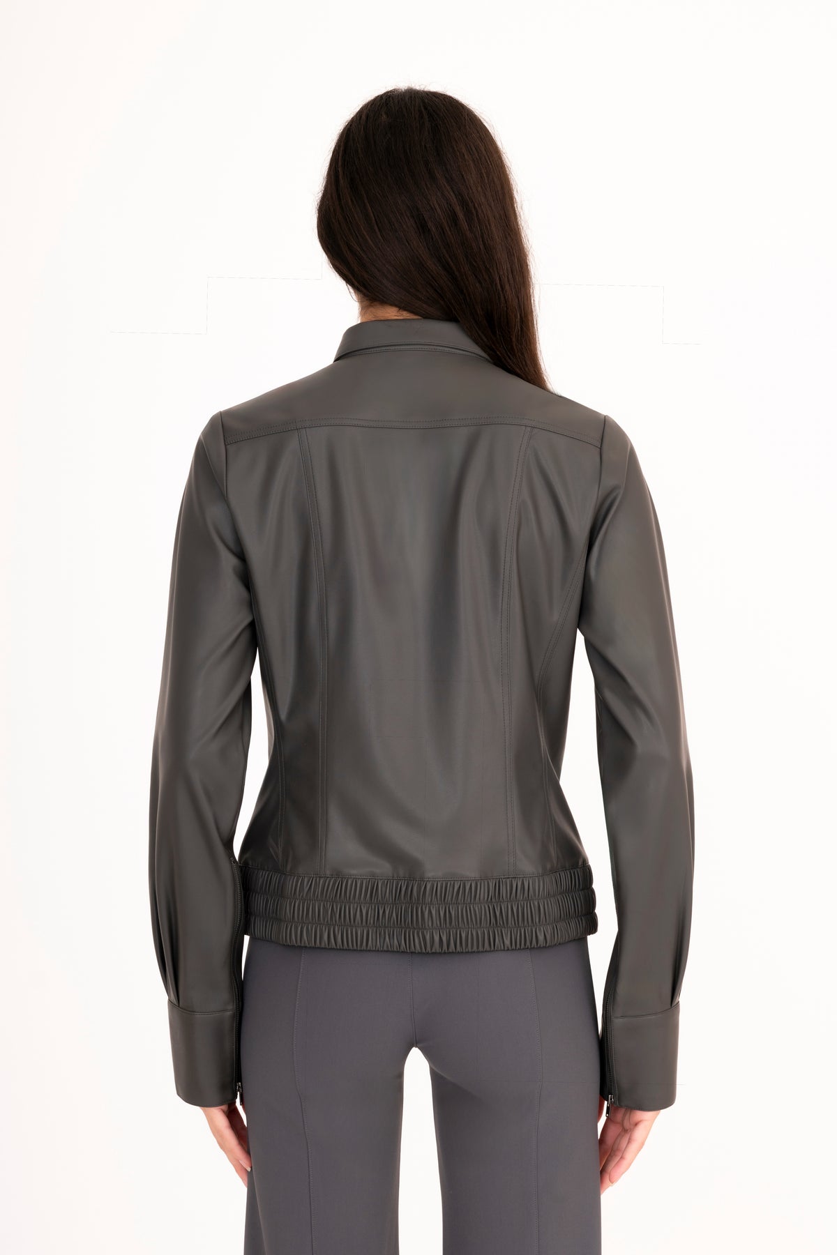 A woman viewed from the back wearing a dark gray vegan leather jacket with a gathered elastic hem and matching gray pants, set against a plain white studio background.