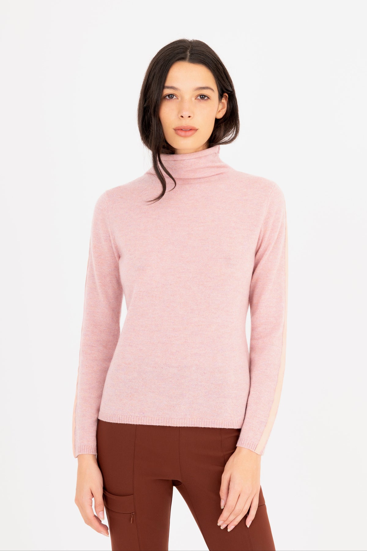 A woman modeling a light pink turtleneck sweater, paired with brown pants against a plain white studio background.
