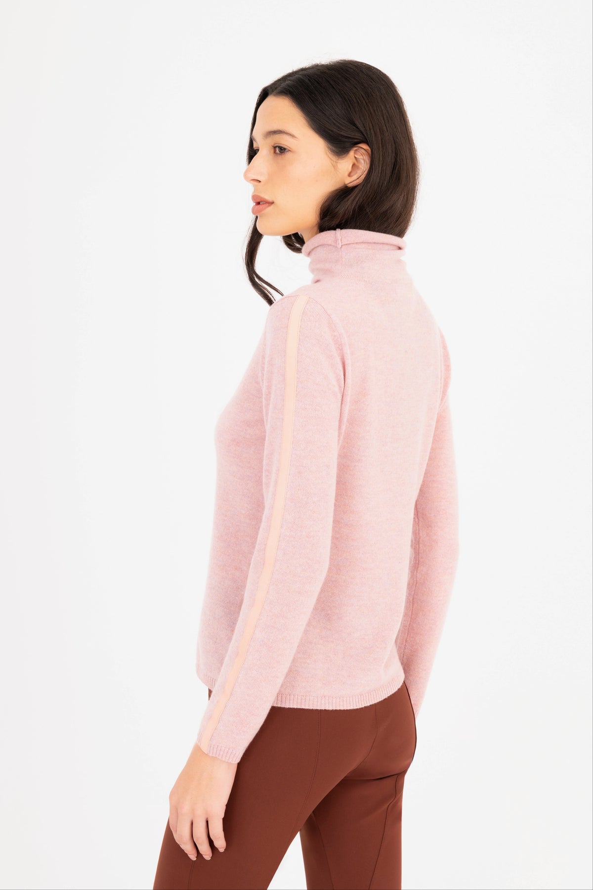 A woman shown from the side profile wearing a light pink mock-neck knit sweater, paired with brown pants, against a plain white background.