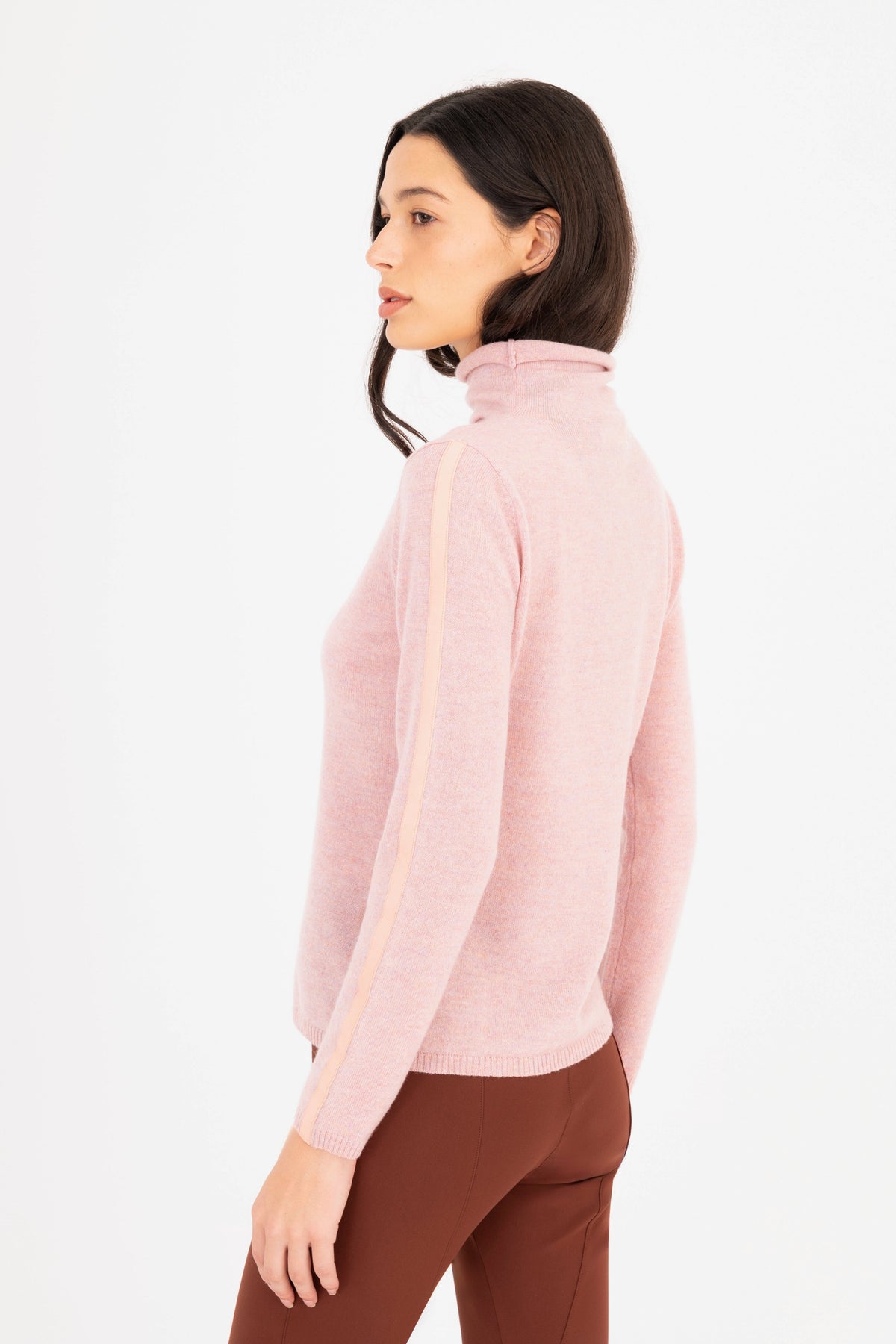 A woman shown from the side profile wearing a light pink mock-neck knit sweater, paired with brown pants, against a plain white background.