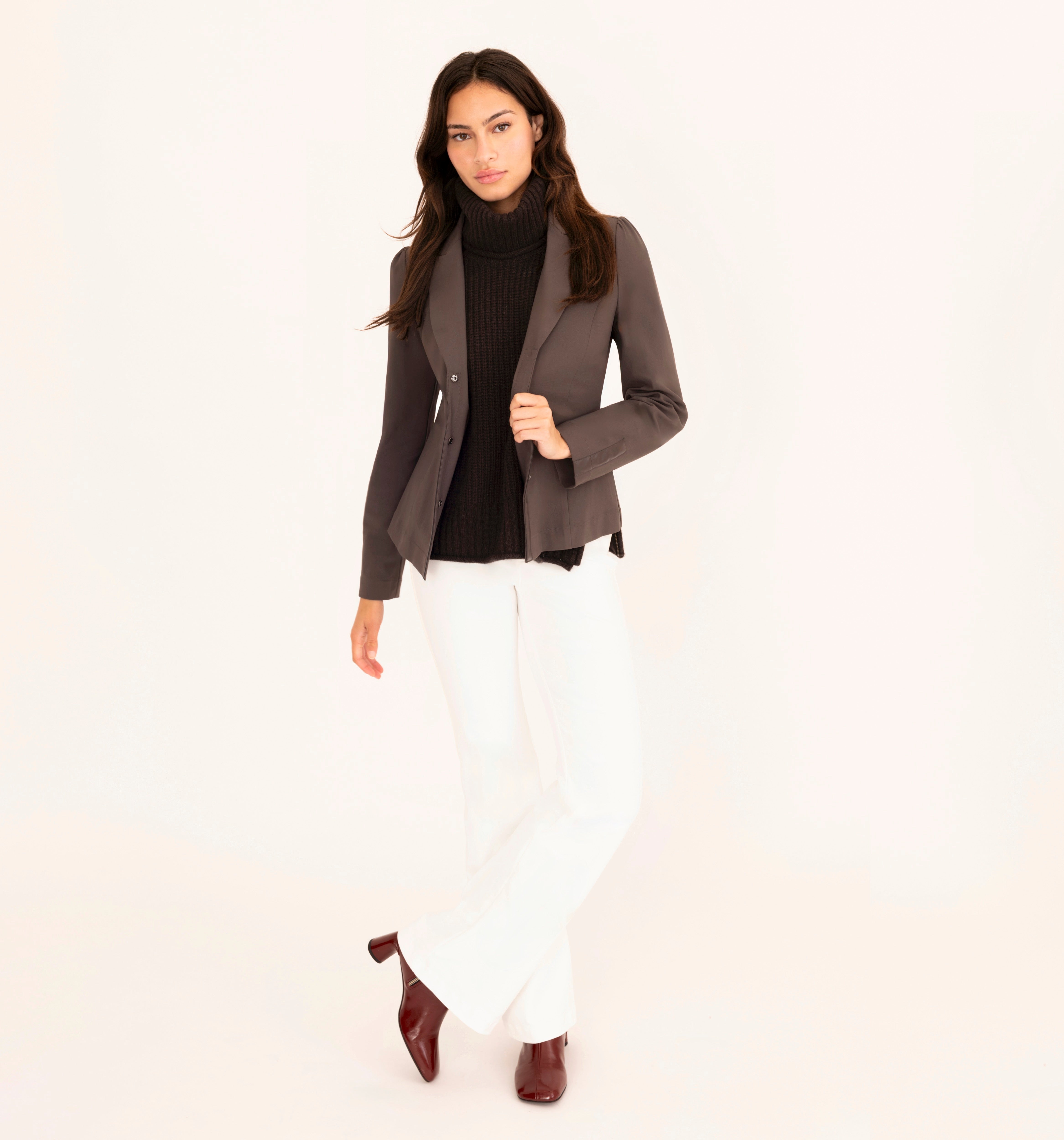 Woman wearing a brown blazer, brown turtleneck, white pants, and brown shoes on a white background