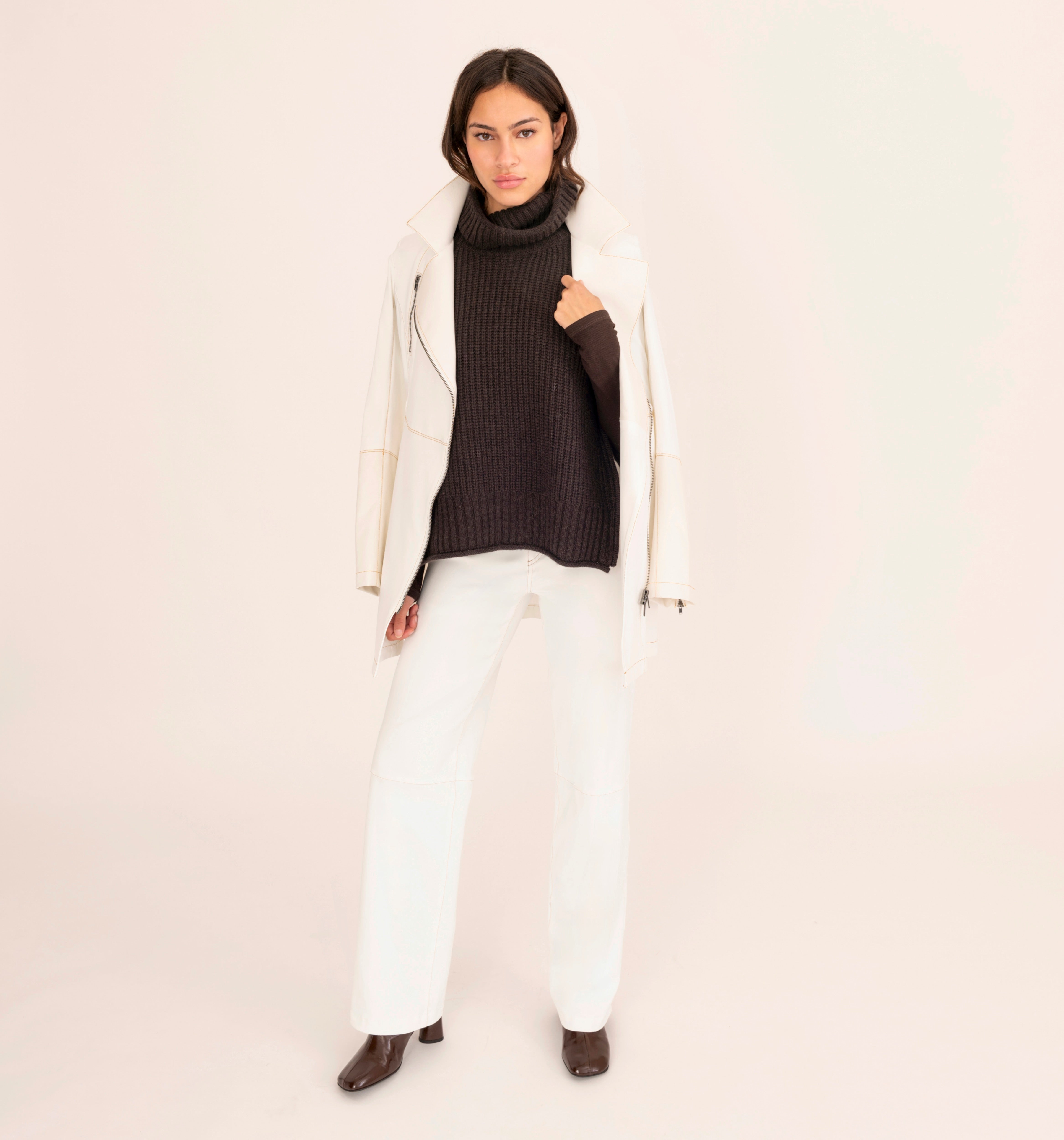 Woman wearing a white coat, brown turtleneck, and white pants on a beige background