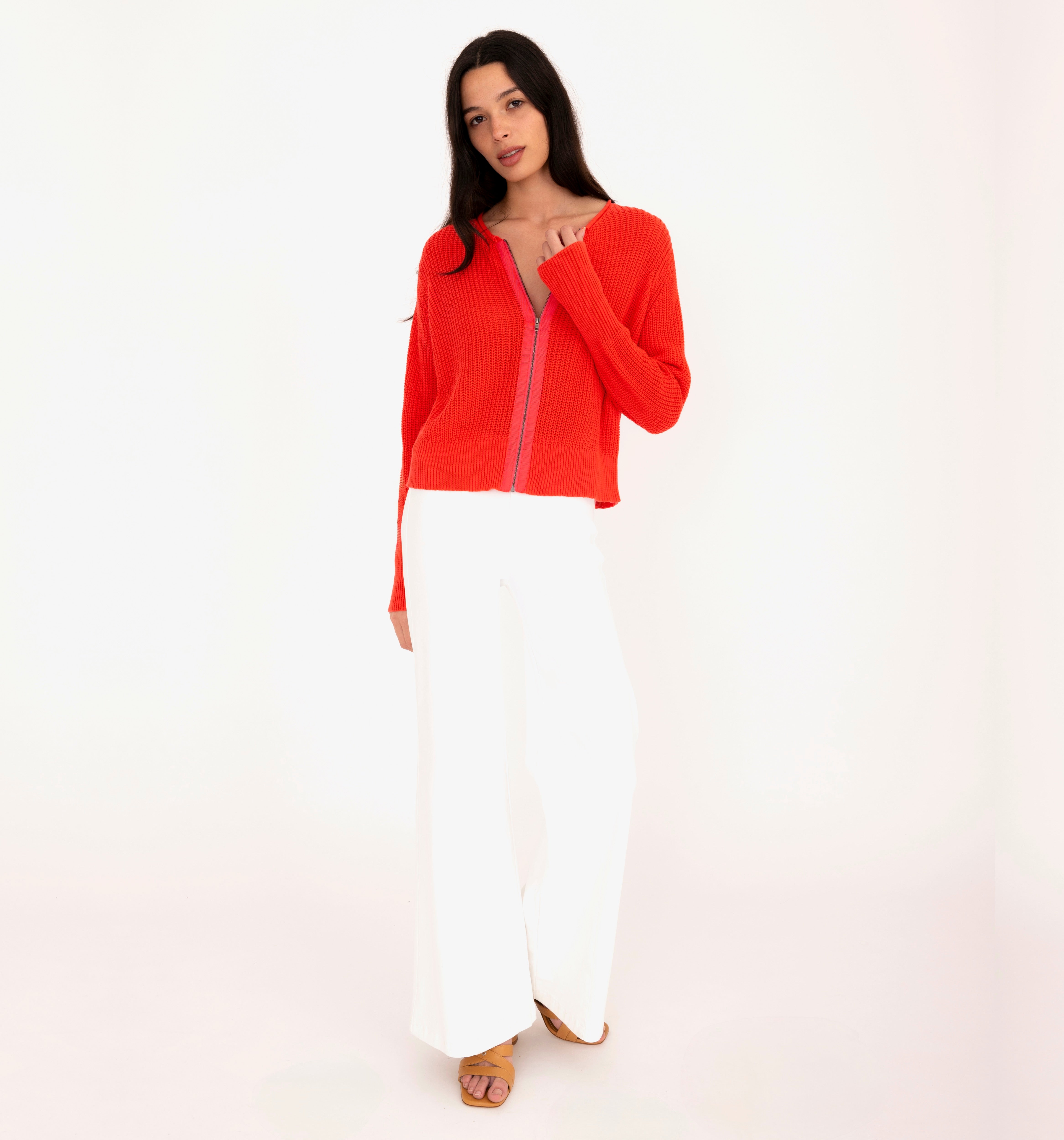 Woman wearing a orange sweater with a zipper and white pants on a white background
